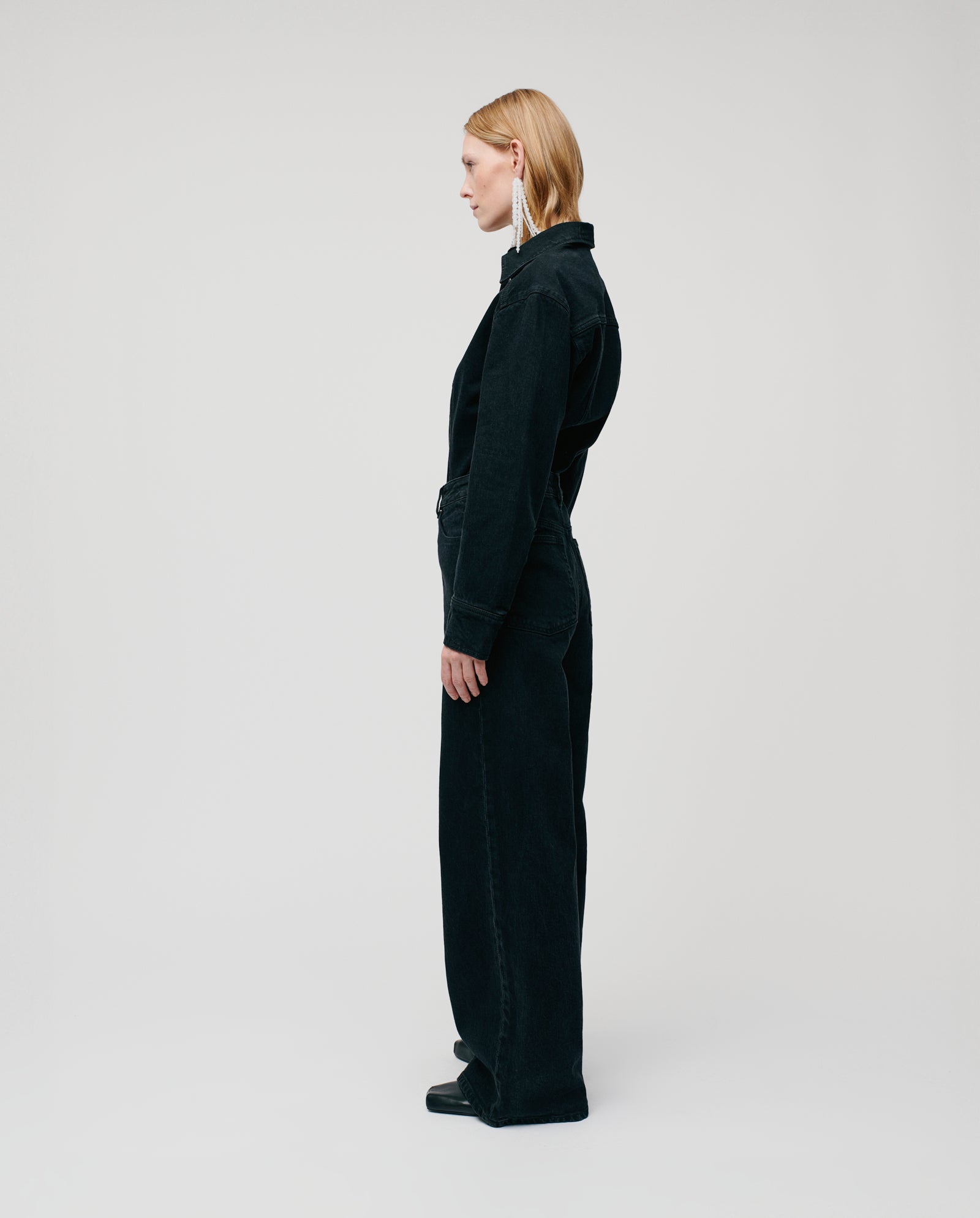 A blond-haired person stands in profile, wearing IVY OAKs PRIJANKA Trousers—high-waisted, loose wide-leg denim—paired with a black long-sleeve top and black shoes.