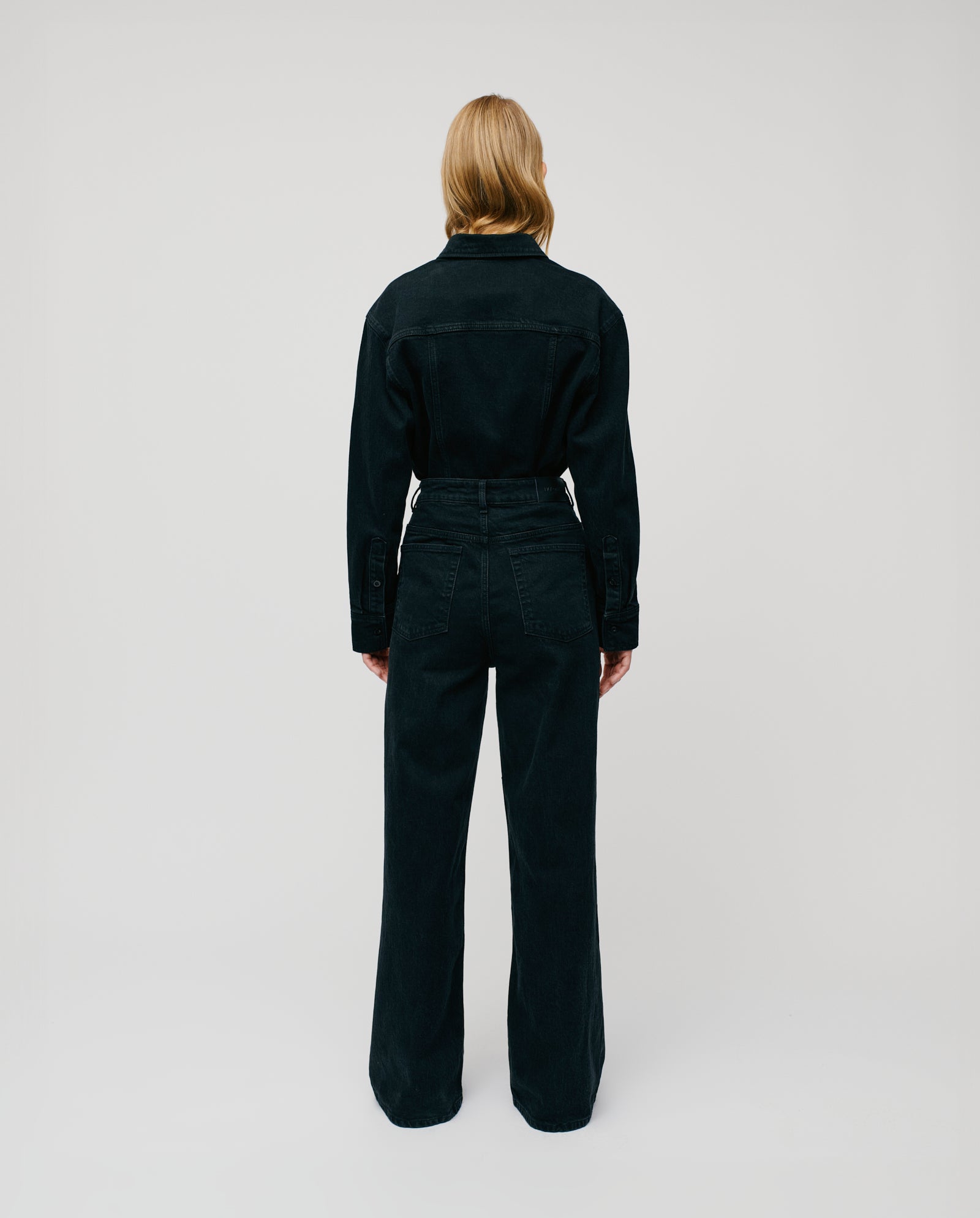 A person with long blonde hair wears the IVY OAK PRIJANKA Trousers and a black denim jacket, standing facing away in a white studio.
