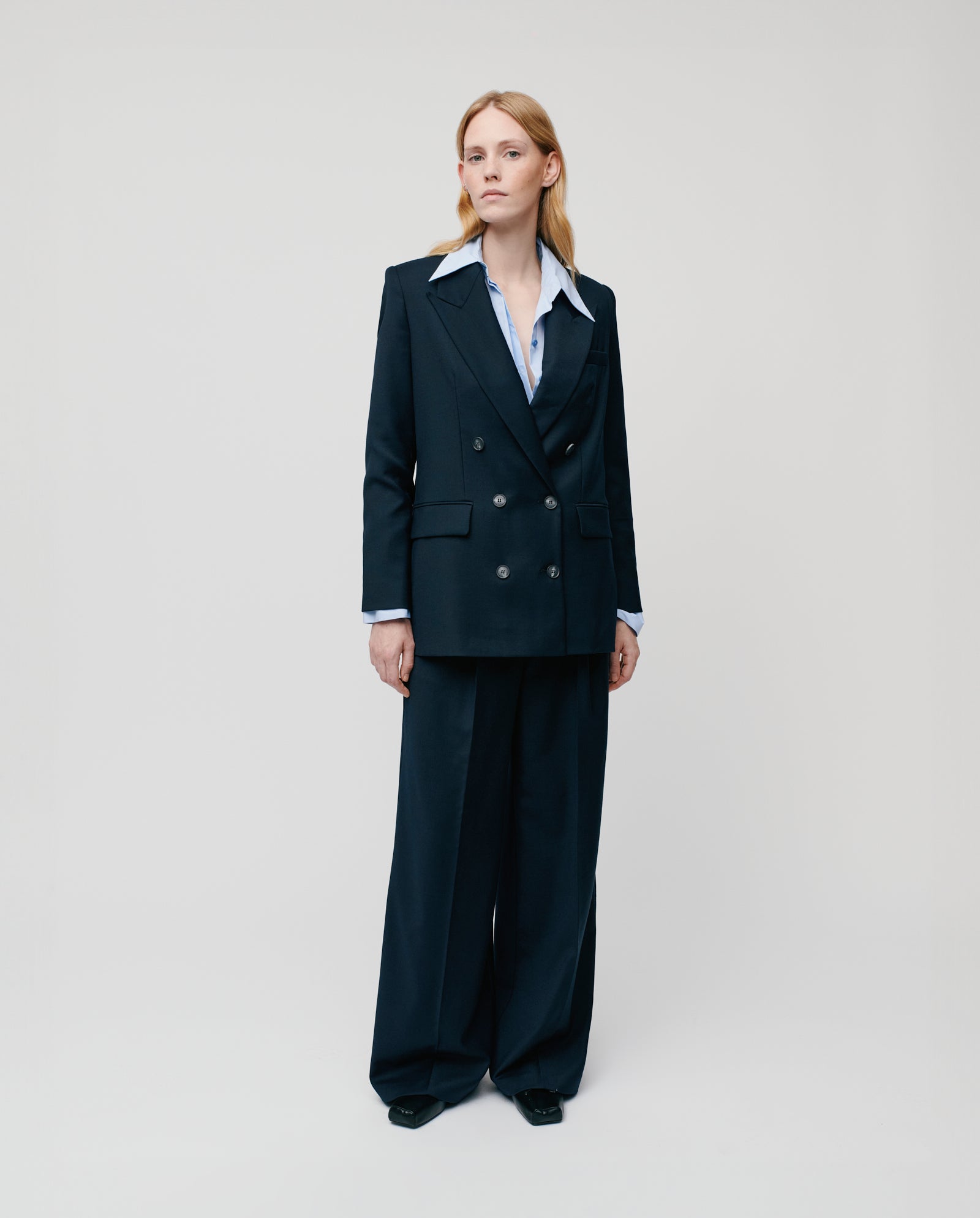 A woman stands against a plain background in the IVY OAK JULIETTE ROSE Blazer, a navy blue double-breasted design paired with wide-leg pants and a white collared shirt, featuring button closure.