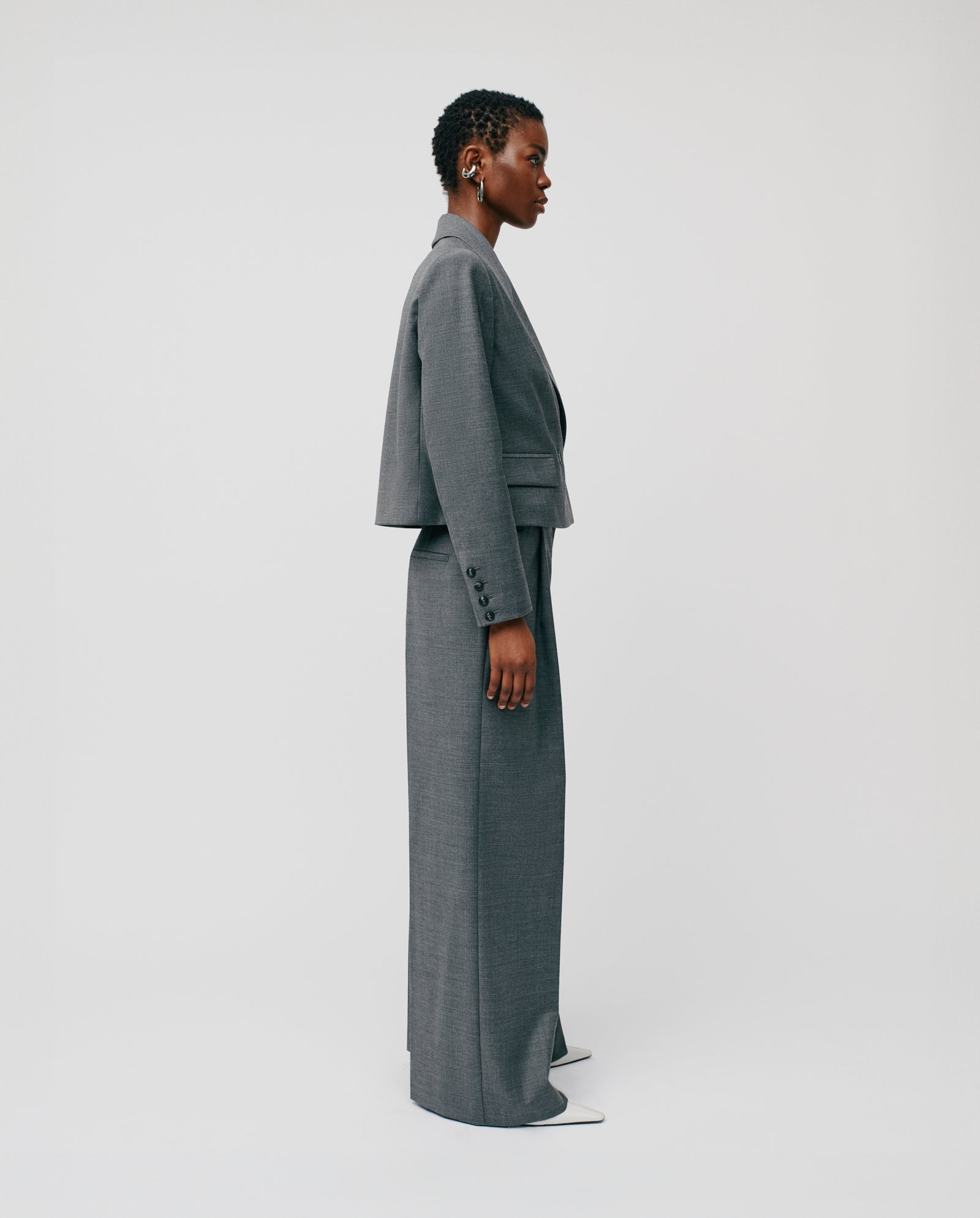 A person with a tall frame wears IVY OAK PRESCILLIA trousers and a JOLANKA blazer, standing in profile against a plain white background.