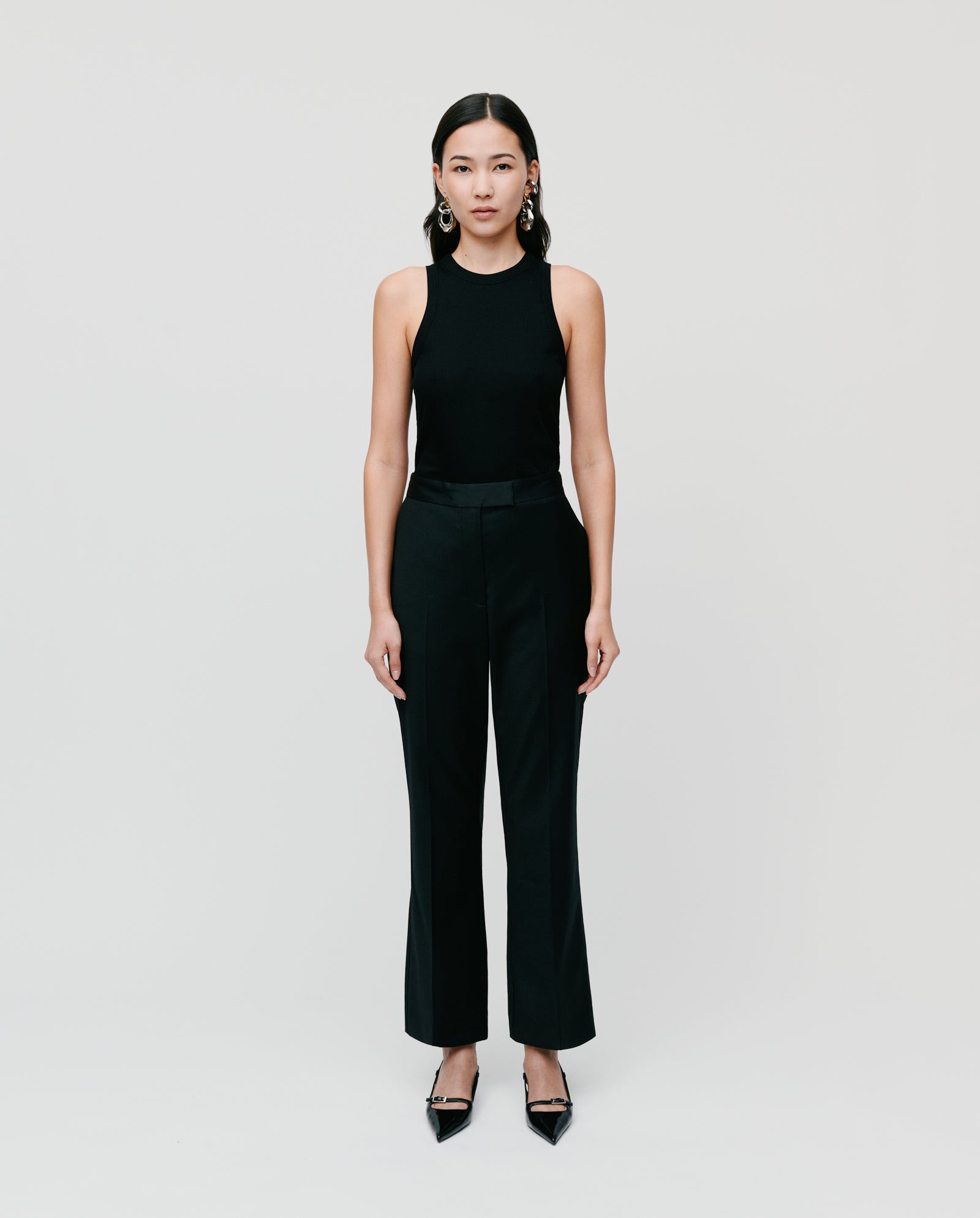 A woman stands against a plain background, wearing IVY OAKs POLINA ROSE Trousers paired with a black sleeveless top and black heels.