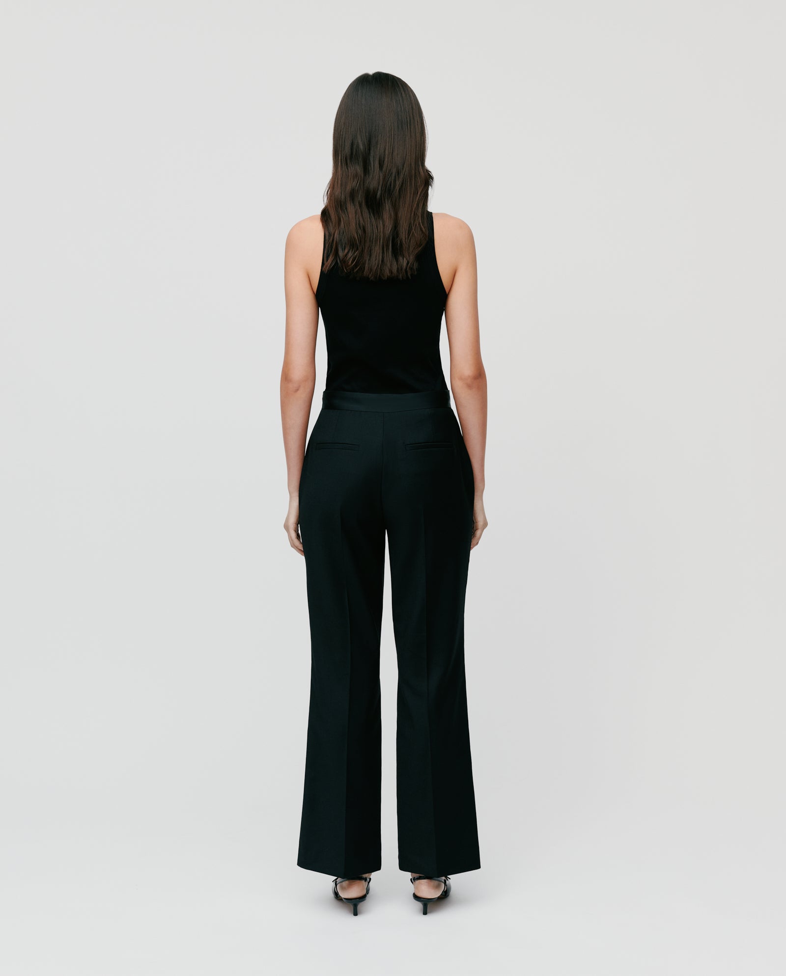 A woman stands facing away on a plain background, wearing IVY OAK’s POLINA ROSE Trousers with a shiny waistband, styled with a black sleeveless top and heels.
