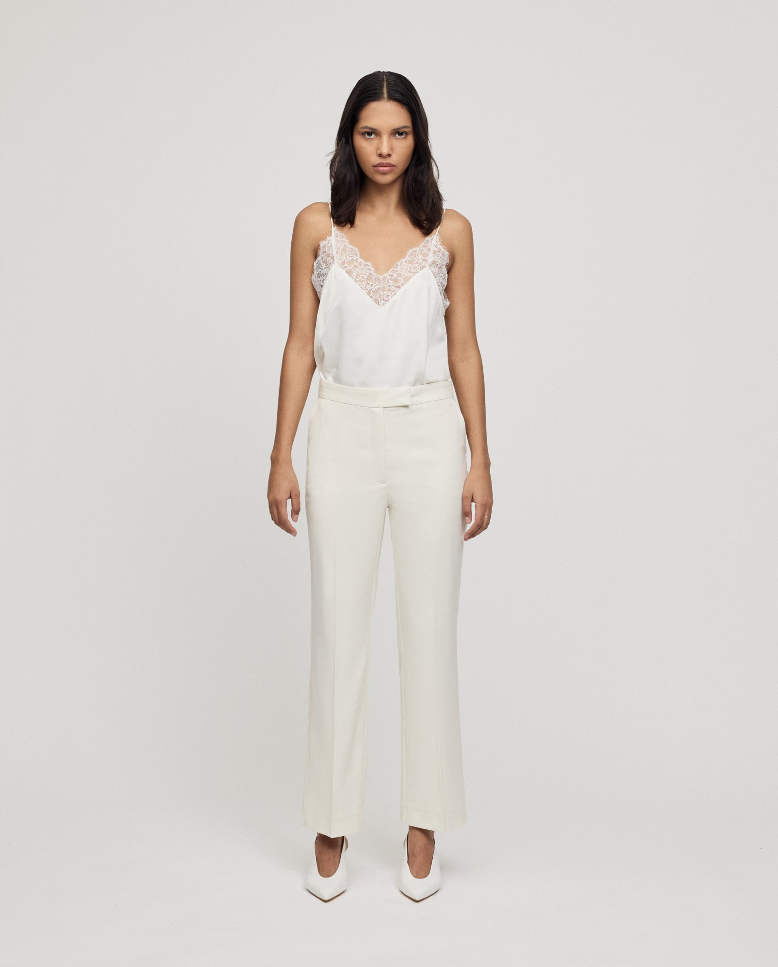 A woman stands against a plain background wearing fitted white POLINA ROSE Trousers by IVY OAK, a lace-trimmed camisole, and white heels.