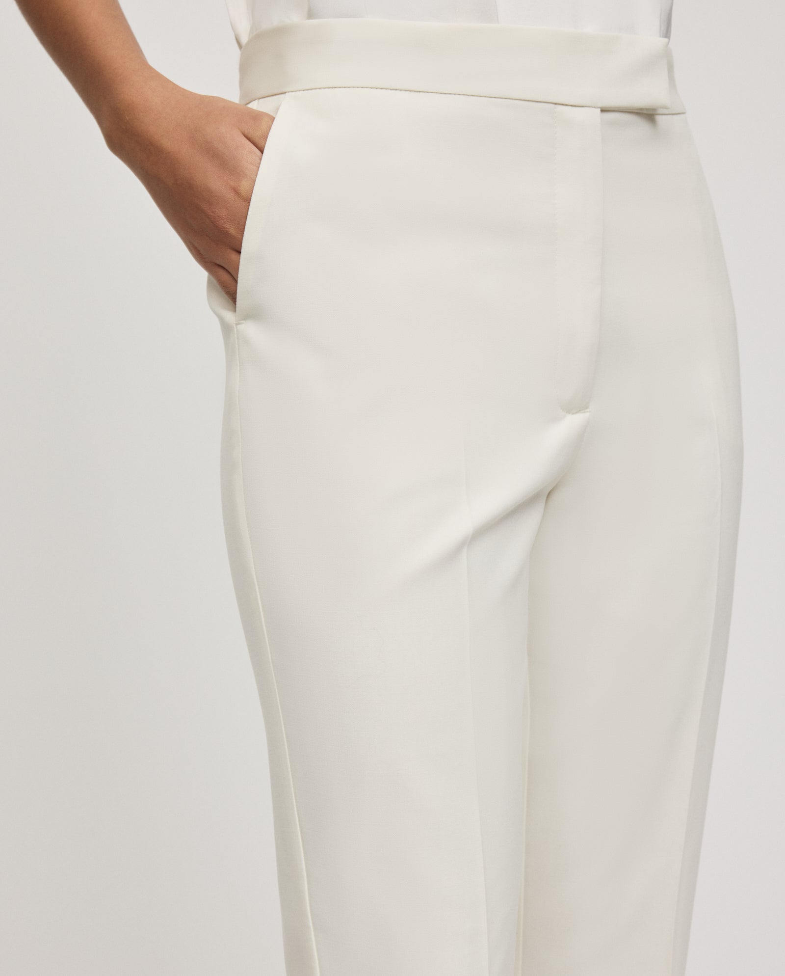 Model wears IVY OAKs POLINA ROSE Trousers—high-waisted, white fitted pants with a sharp crease and hand in pocket—set against a plain background.