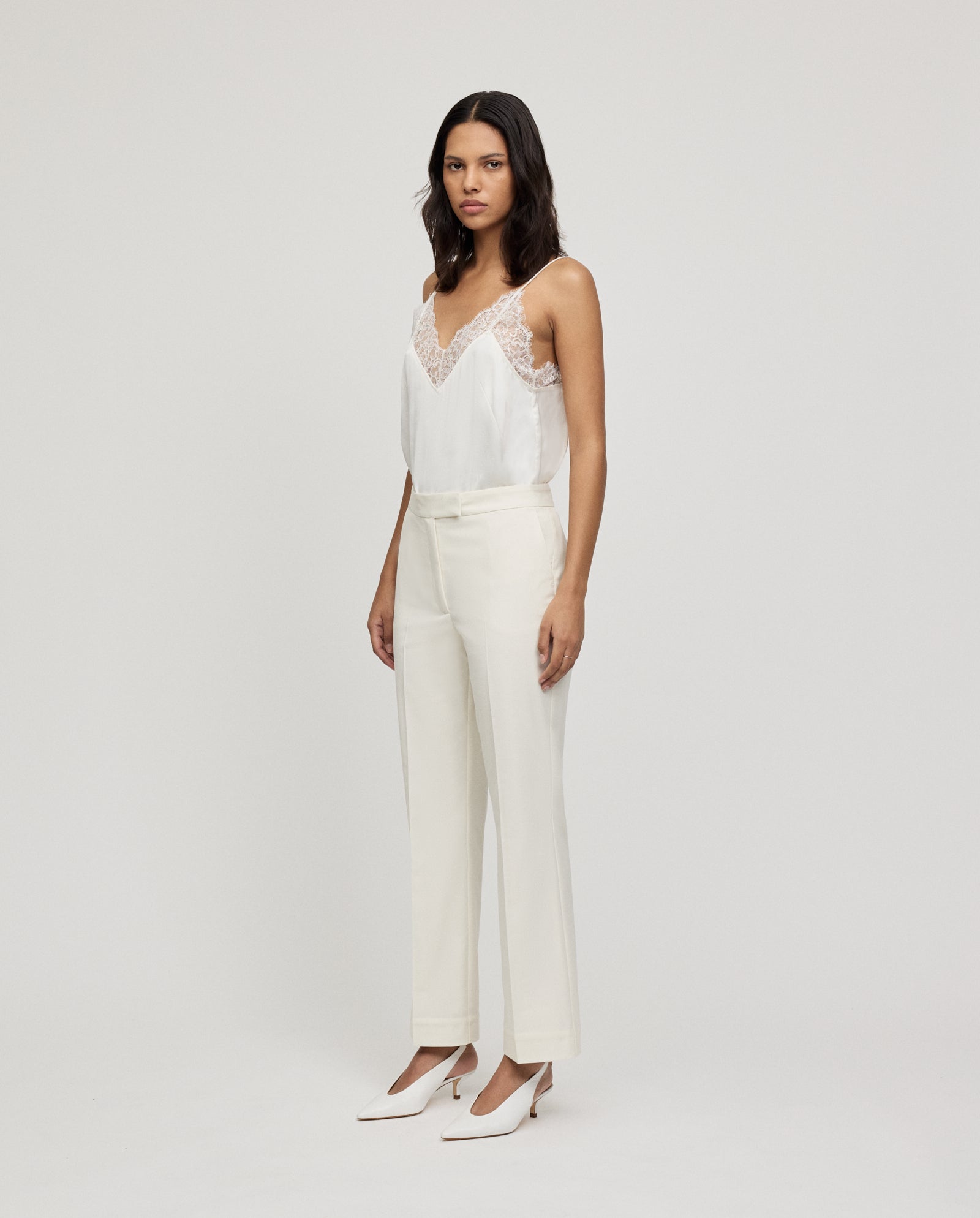 A woman in a white lace-trim camisole, POLINA ROSE Trousers by IVY OAK, and white heels stands against a plain background.