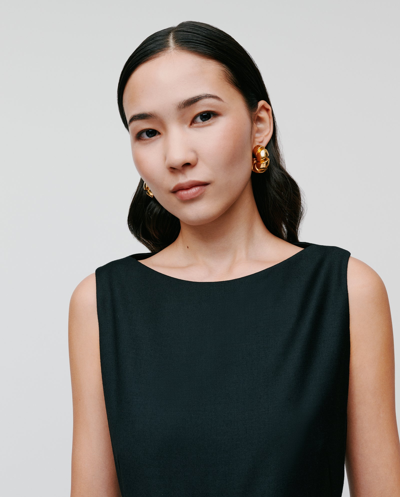 Woman with long dark hair wears the IVY OAK PEPPINA Jumpsuit, a sleeveless black piece with a boat neckline, paired with large gold earrings, set against a plain background.