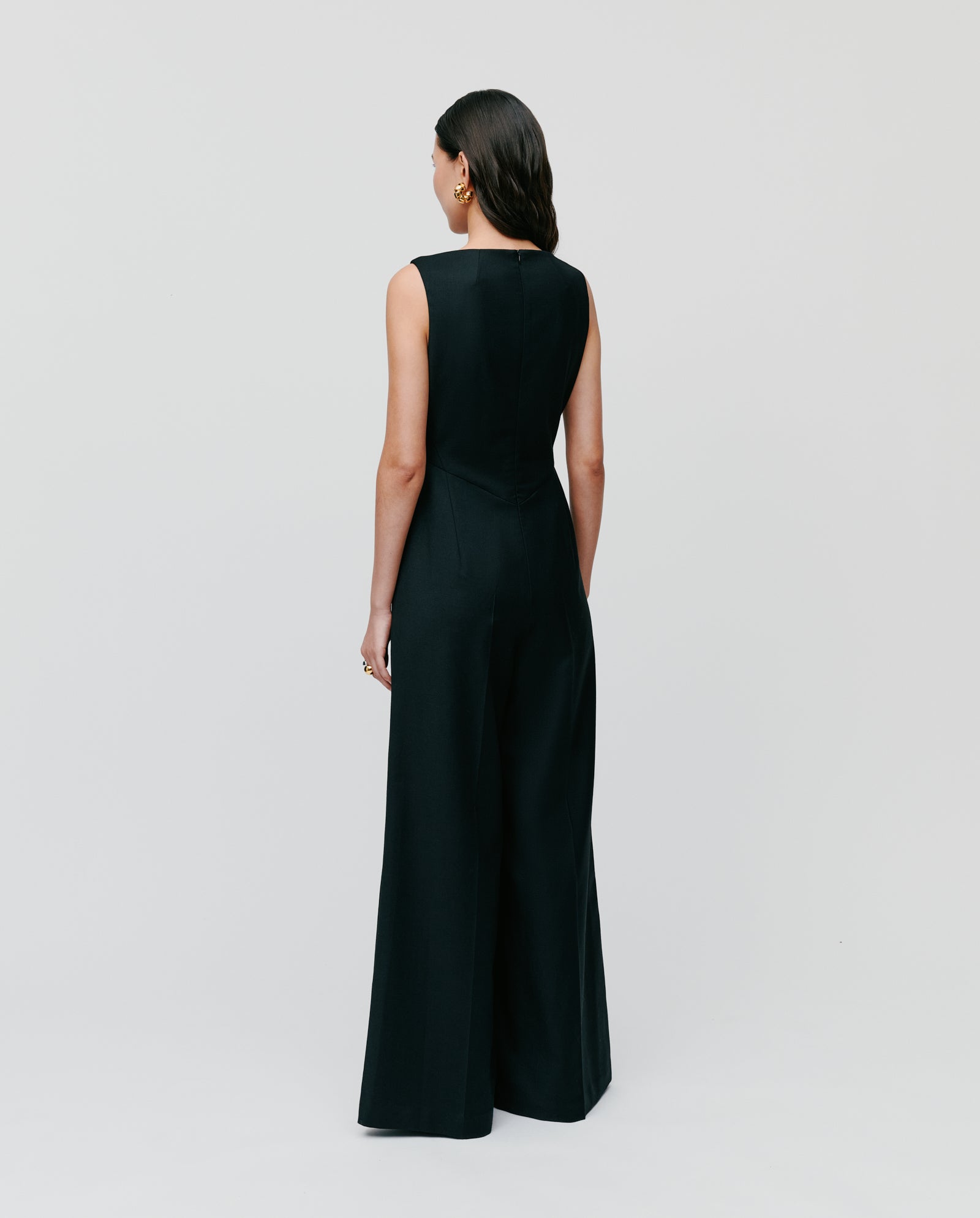 A woman with long dark hair stands facing away from the camera, wearing the sleeveless black PEPPINA Jumpsuit by IVY OAK, featuring wide legs.