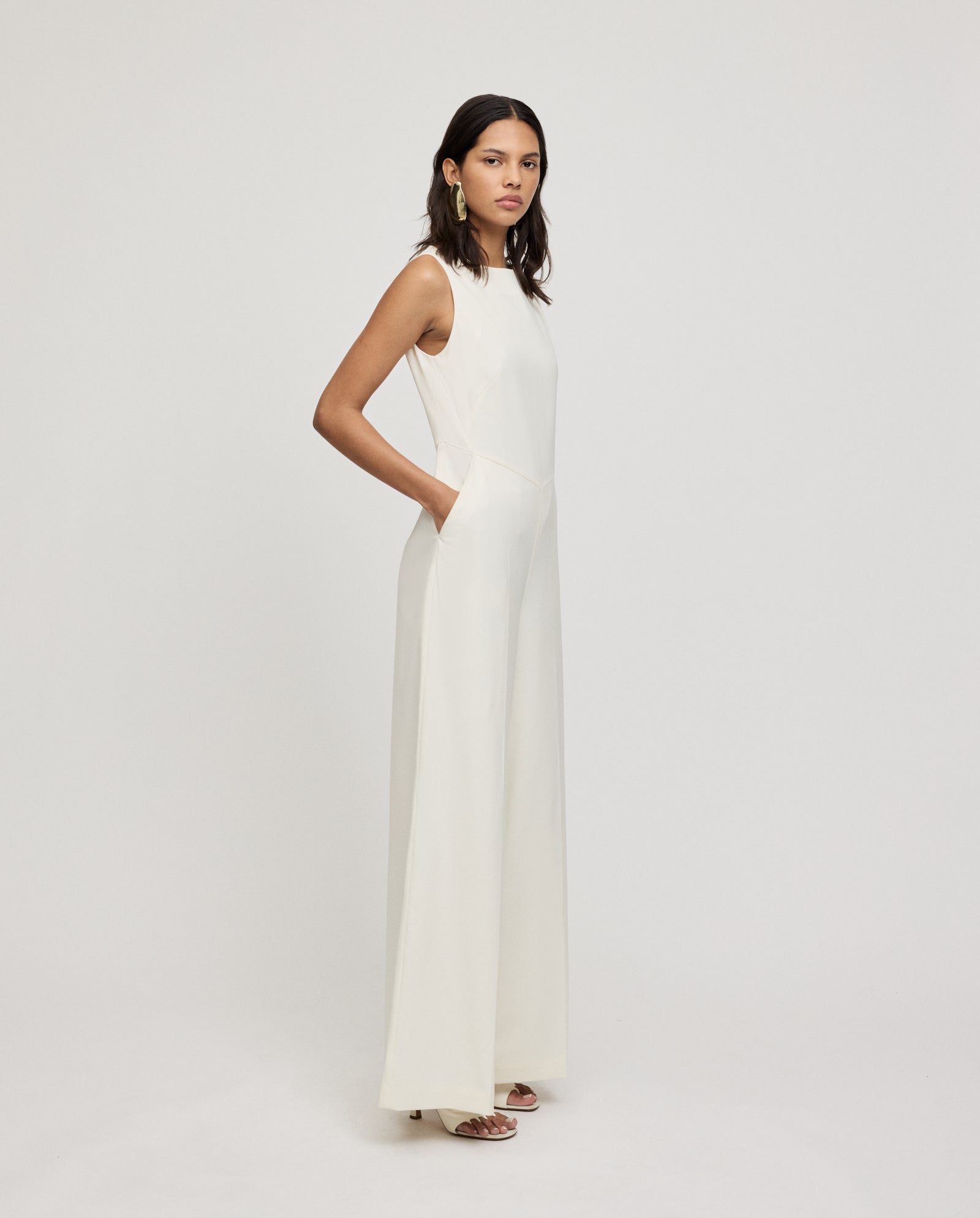 A woman wears the IVY OAK PEPPINA Jumpsuit, a sleeveless white design with a boat neckline and wide-leg pants, standing against a plain white background with her hands in her pockets.