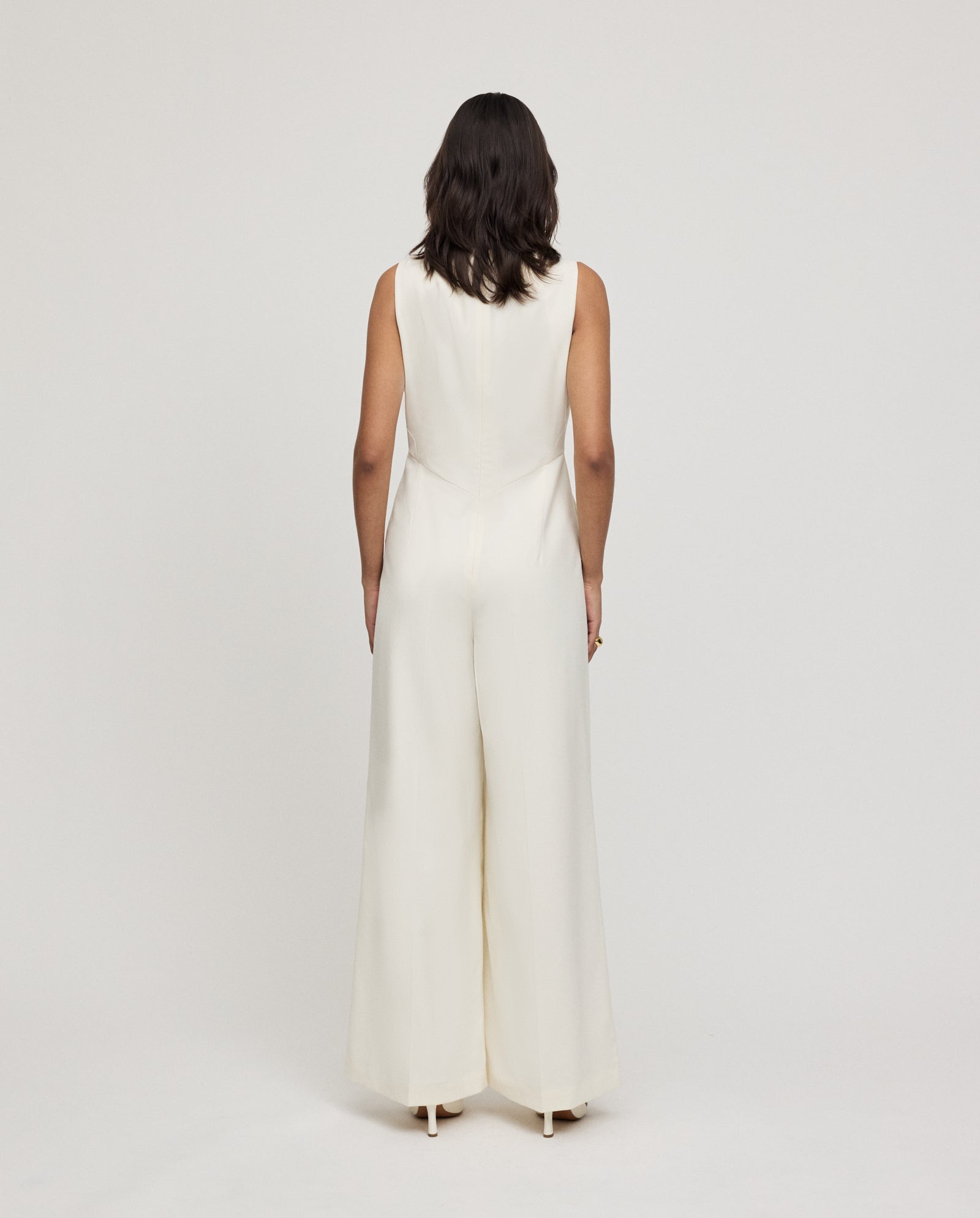 A woman stands facing away against a plain light background, wearing the IVY OAK PEPPINA Jumpsuit—a sleeveless white piece with a boat neckline.