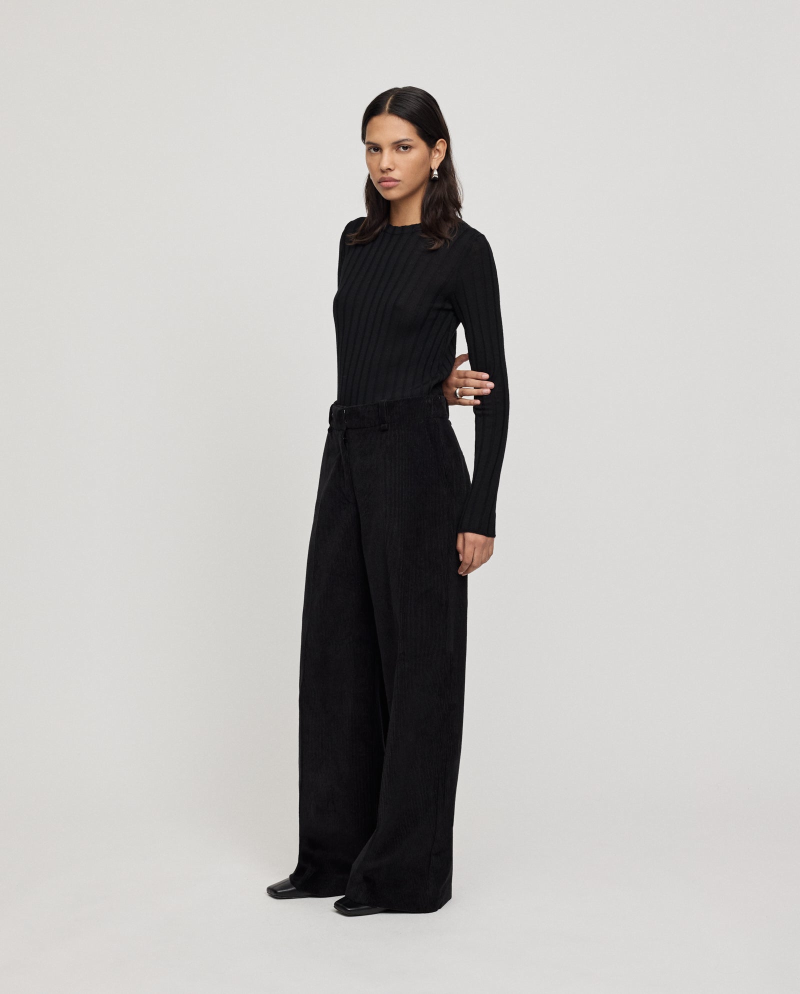 A woman stands against a plain background wearing IVY OAKs PERNILLE ANN high-waisted, wide-leg corduroy trousers paired with a fitted black long-sleeve top.
