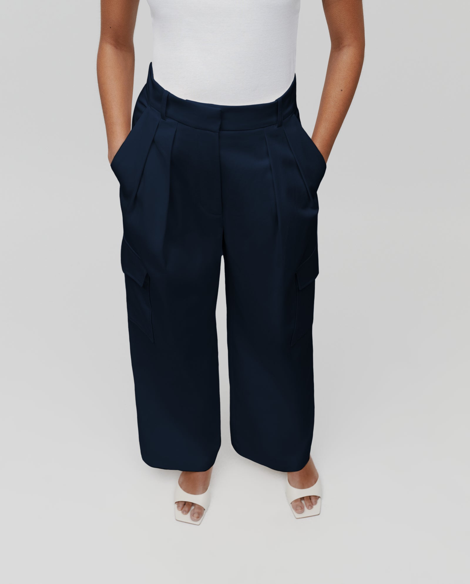 A person wears IVY OAK PRESTIGE Pants in dark blue—barrel leg, fitted waist—paired with a white top and white open-toe heels, hands in pockets.