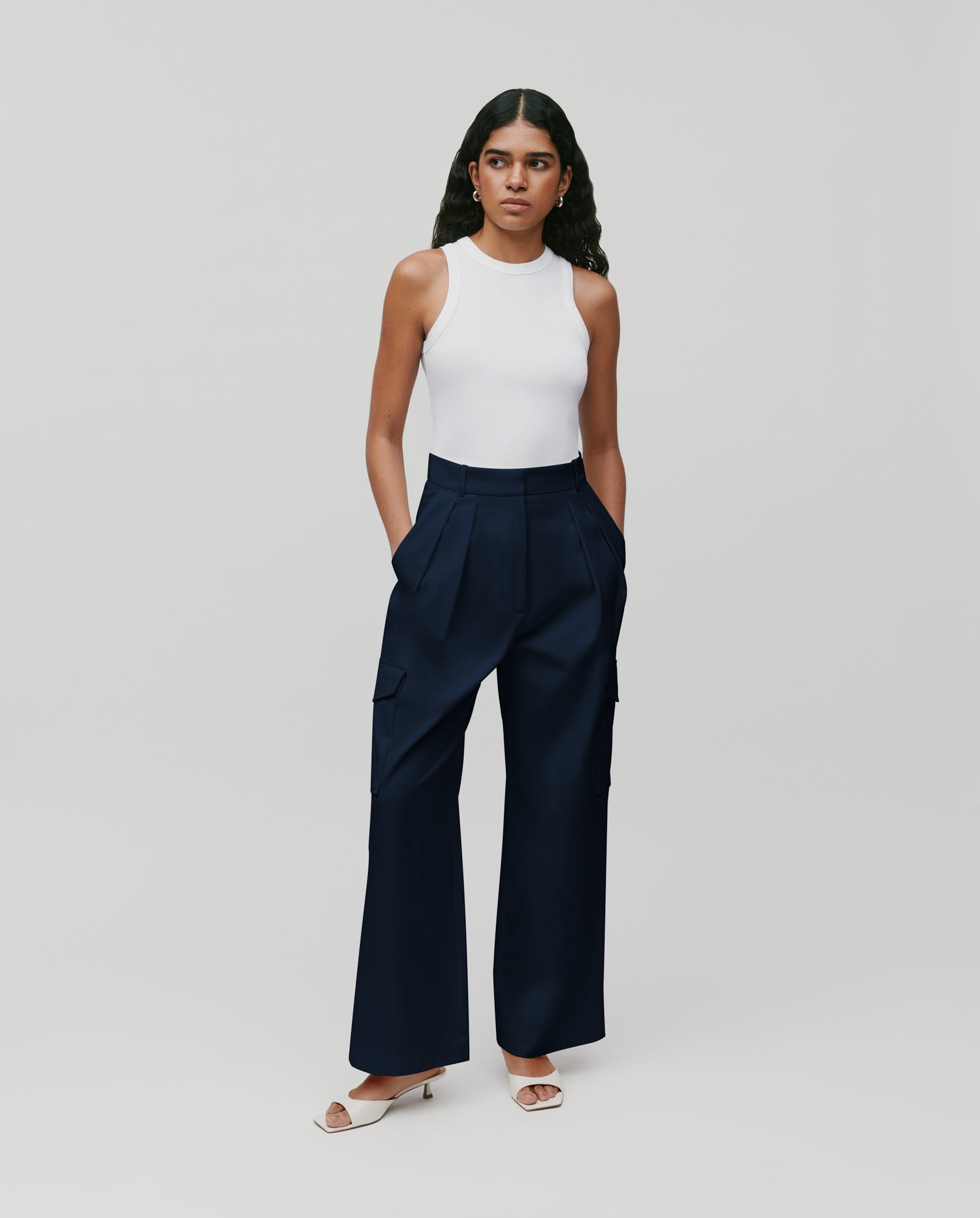 A woman stands against a plain background, radiating effortless style in IVY OAK’s PRESTIGE Pants—navy barrel leg cargo pants with a fitted waist—paired with a white sleeveless top and white heels.