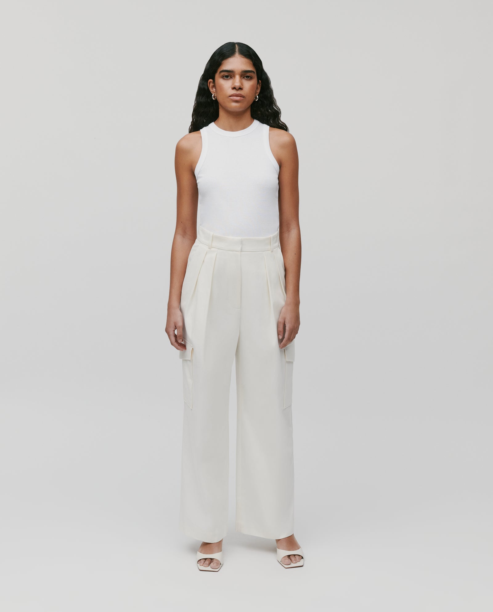 A woman in IVY OAKs PRESTIGE Pants, paired with a white sleeveless top and sandals, stands against a plain light background.