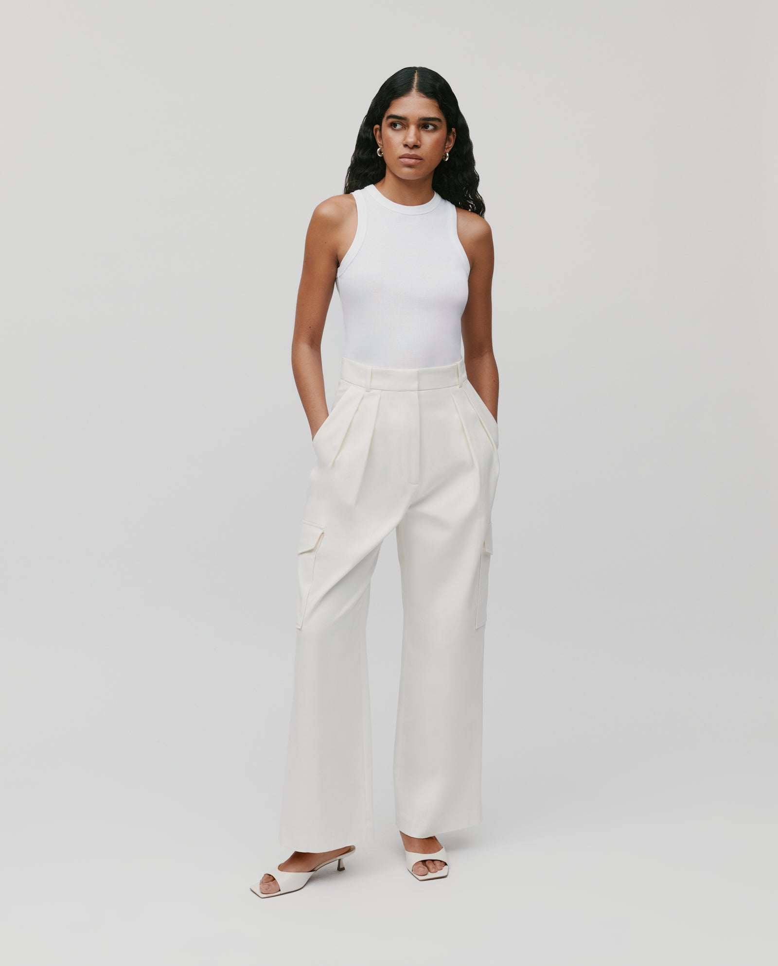 A woman stands against a plain background wearing IVY OAKs white PRESTIGE Pants with a fitted waist, paired with a white sleeveless top and white sandals.