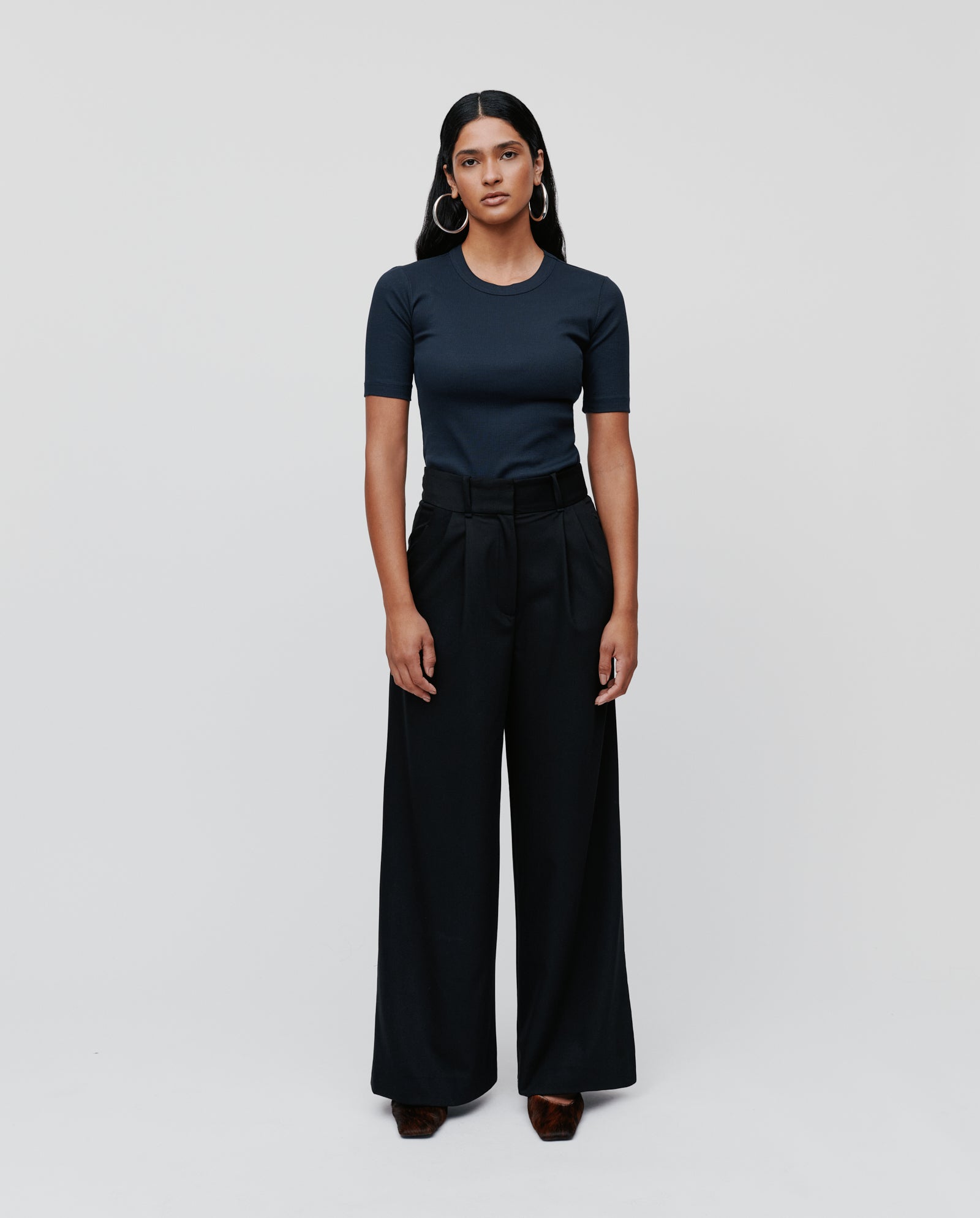 A woman stands against a plain background wearing the IVY OAK PRESCILLIA ANN Trousers, styled with French pockets, paired with a navy t-shirt.