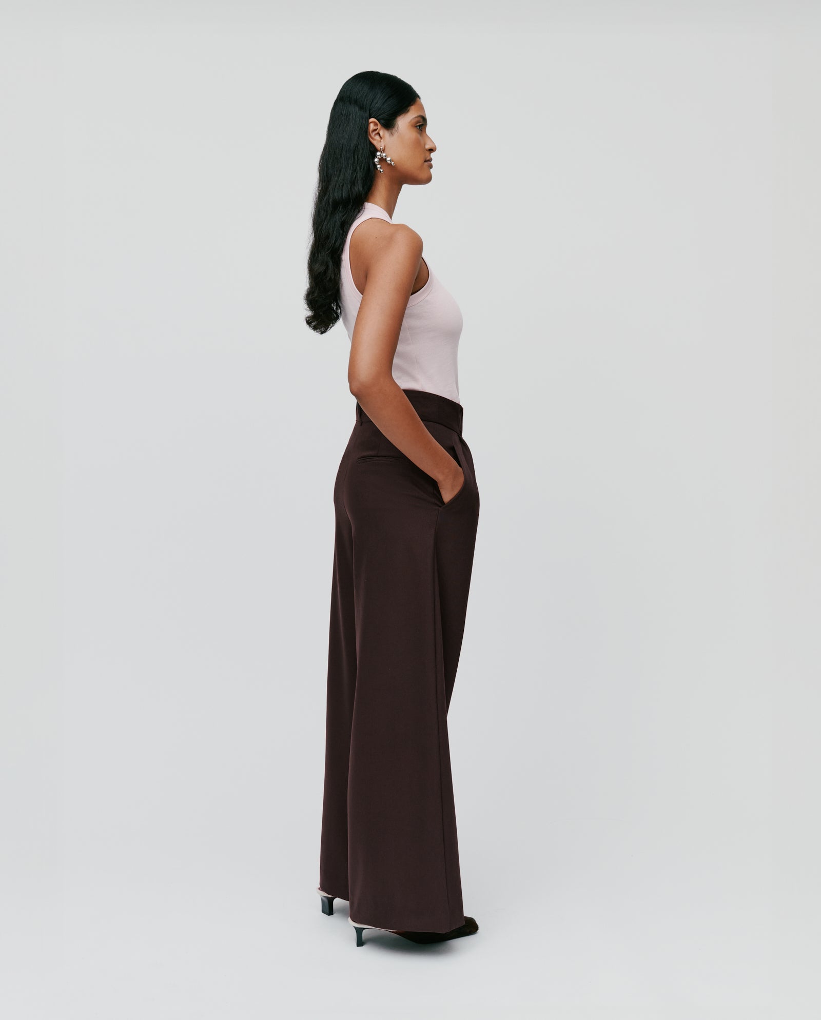 A woman stands sideways with her hands in the pockets of IVY OAK’s PRESCILLIA ANN brown high-waist chic trousers with French pockets, paired with a pink sleeveless top.