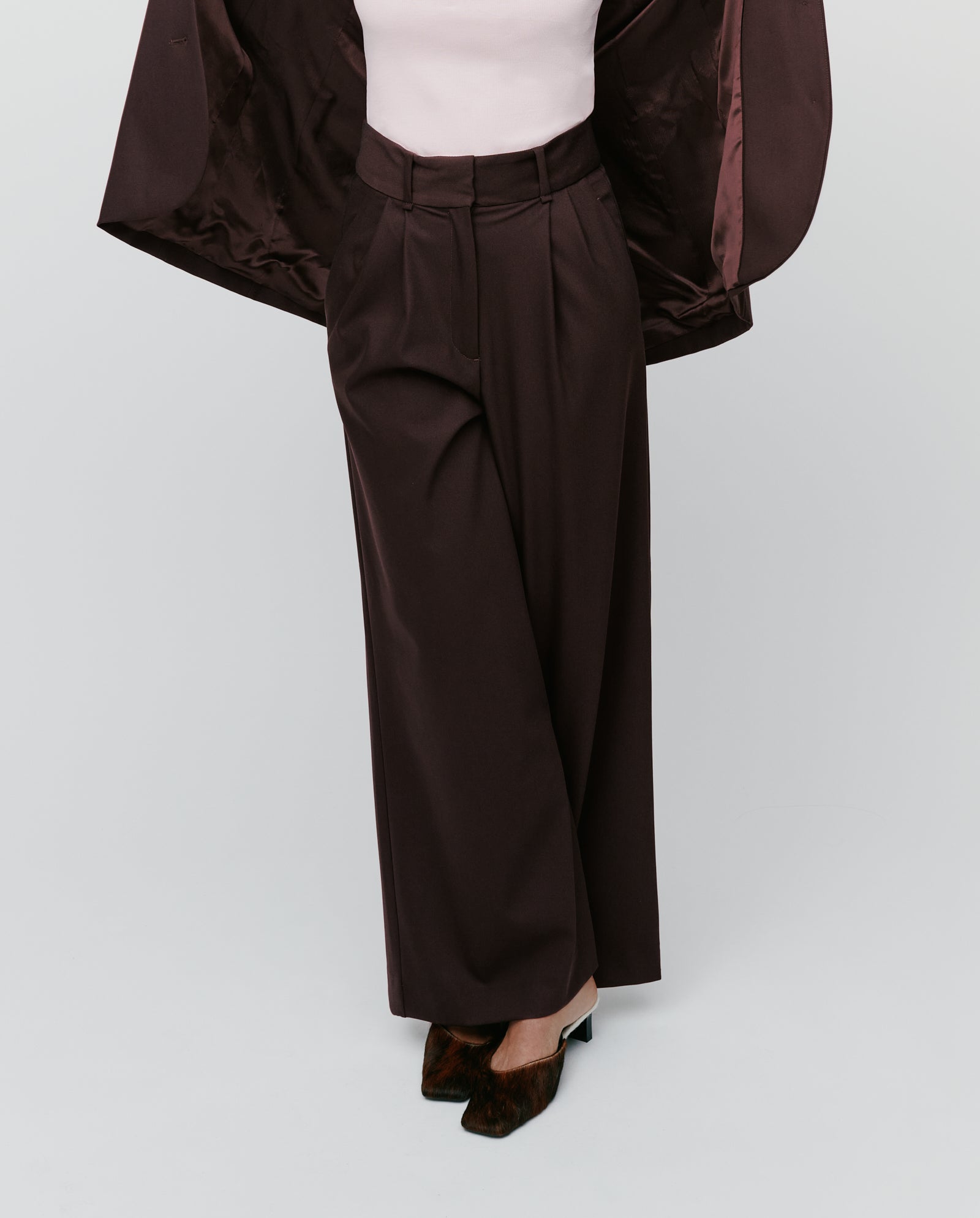 Model wears IVY OAKs PRESCILLIA ANN high-waist, wide-leg brown trousers with French pockets, paired with a blush top and brown coat; shown from the shoulders down.