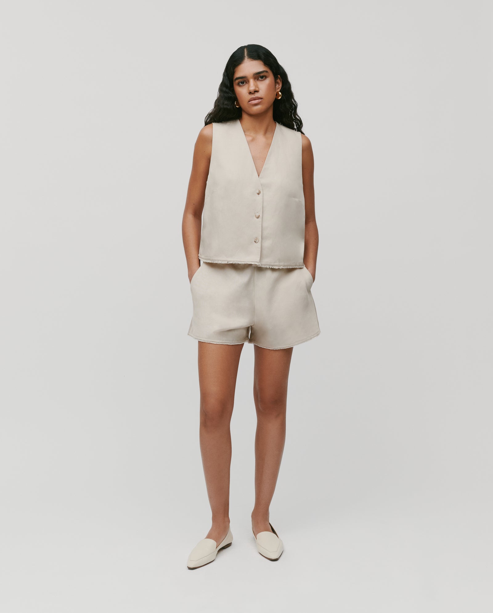 Model wearing IVY OAKs PIPER Trousers—beige, sleeveless, with an elastic waistband—standing hands-in-pockets against a plain background.