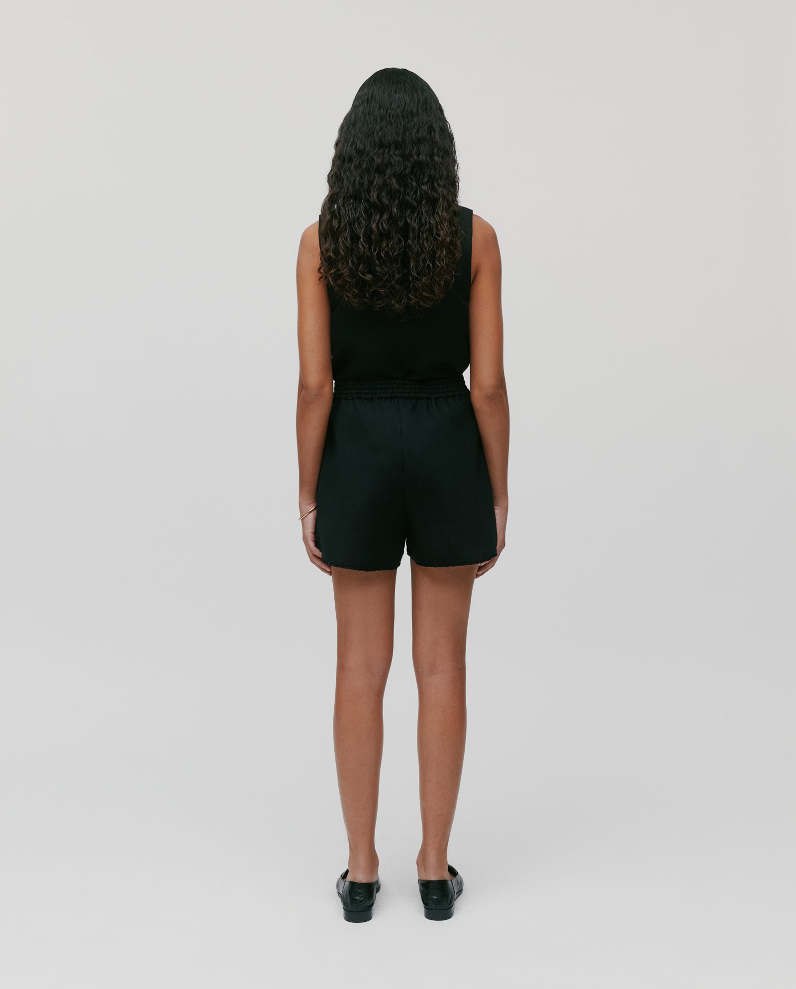 A person with long curly hair, wearing a black sleeveless top and IVY OAKs PIPER Trousers with an elastic waistband, stands facing away against a plain background.