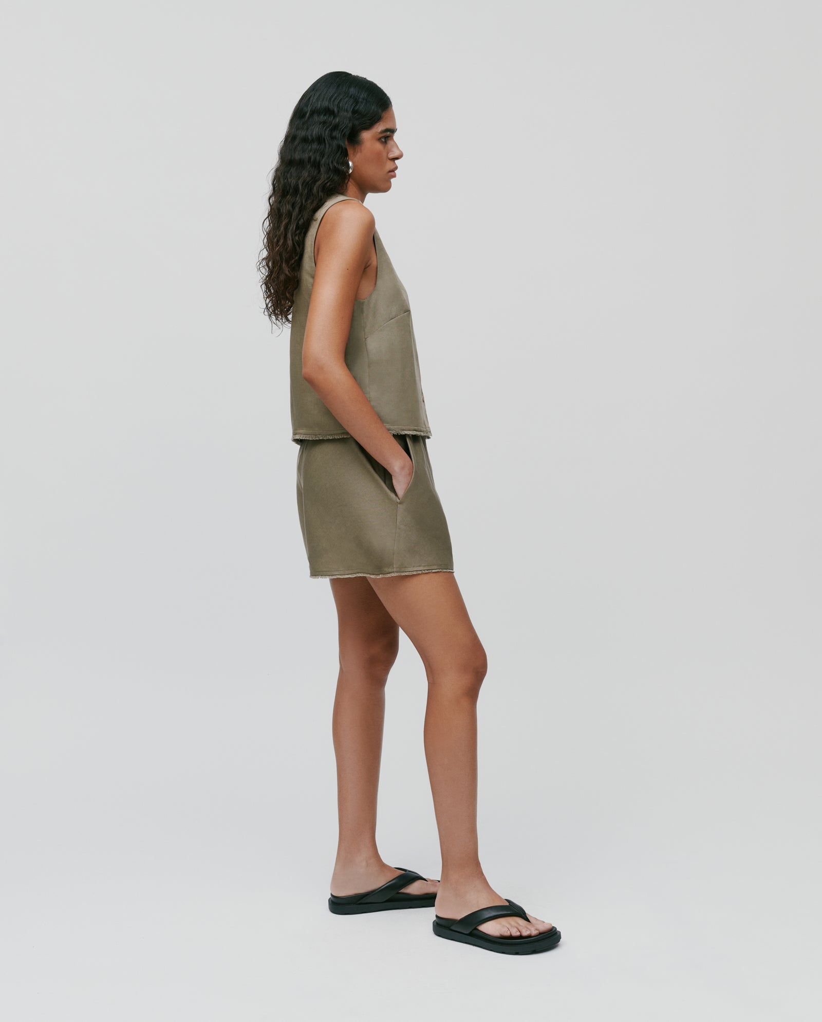 Woman in PIPER Trousers by IVY OAK, olive green sleeveless top, and linen mix shorts with elastic waistband, standing sideways with hands in pockets, wearing black sandals.