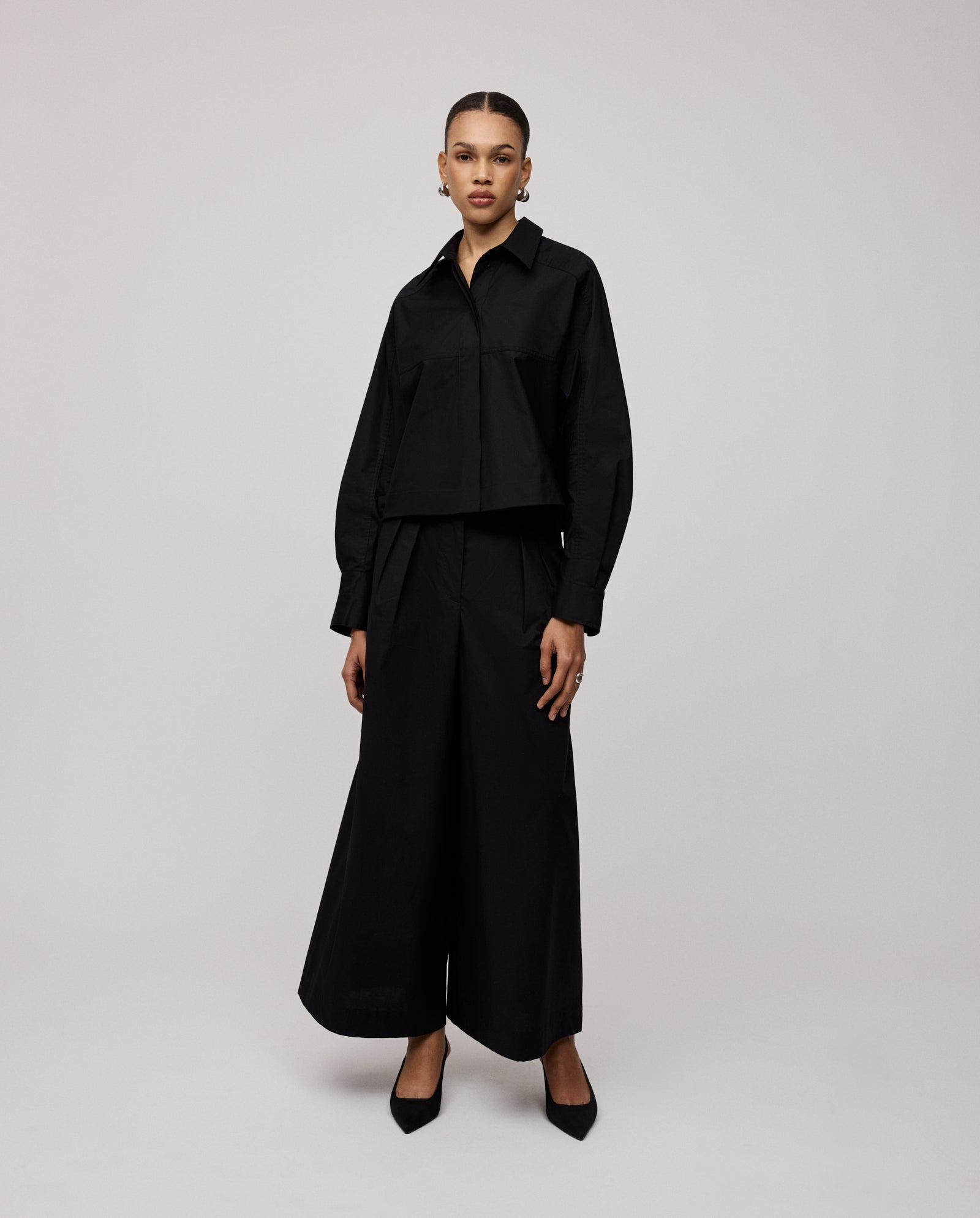 A woman wears IVY OAK’s PIERINA Pants—wide-leg cotton trousers with waist darts—paired with a black long-sleeve top and black heels, standing against a plain background.
