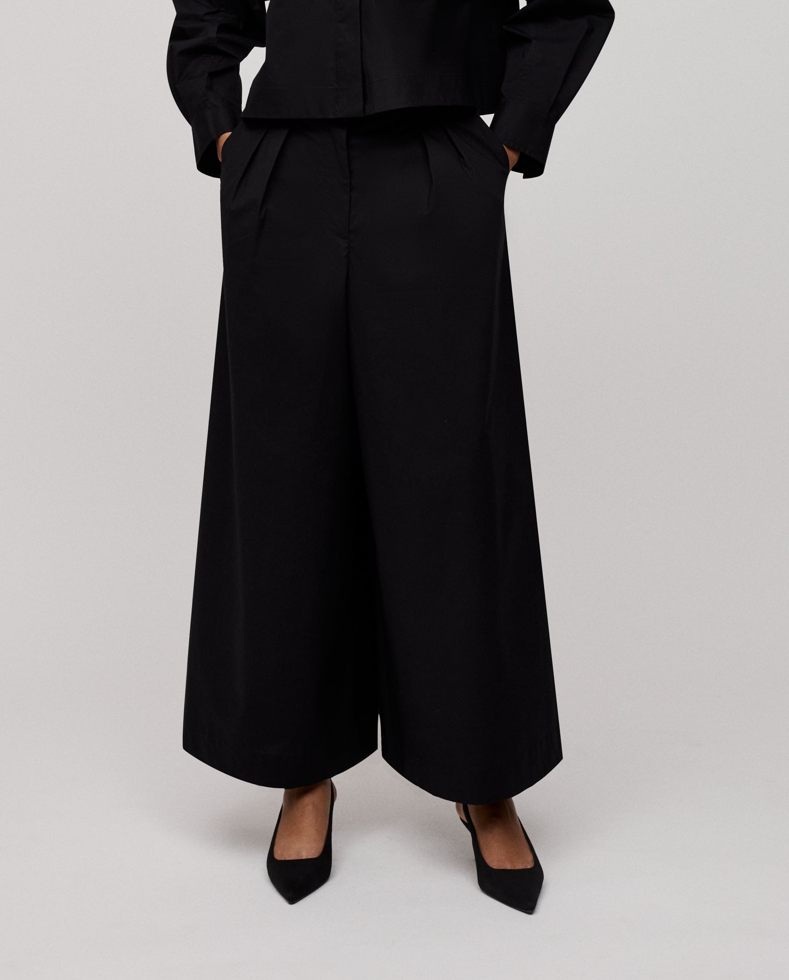 Person wearing IVY OAK PIERINA Pants featuring wide-leg cotton design and waist darts, paired with black heels, hands in pockets, against a plain background.