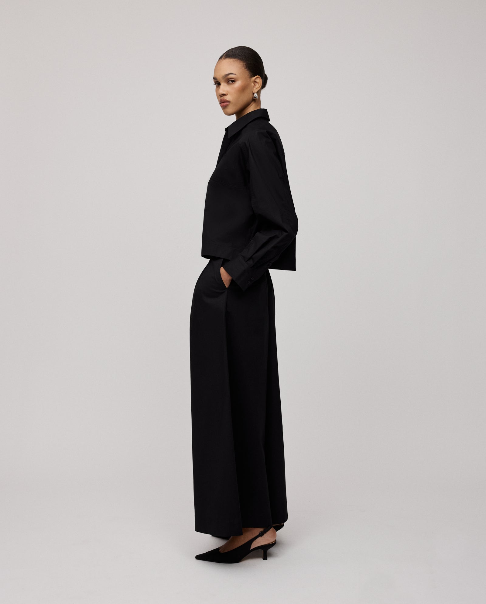 A woman stands sideways with hands in pockets, wearing IVY OAKs PIERINA Pants and a black long-sleeve top.