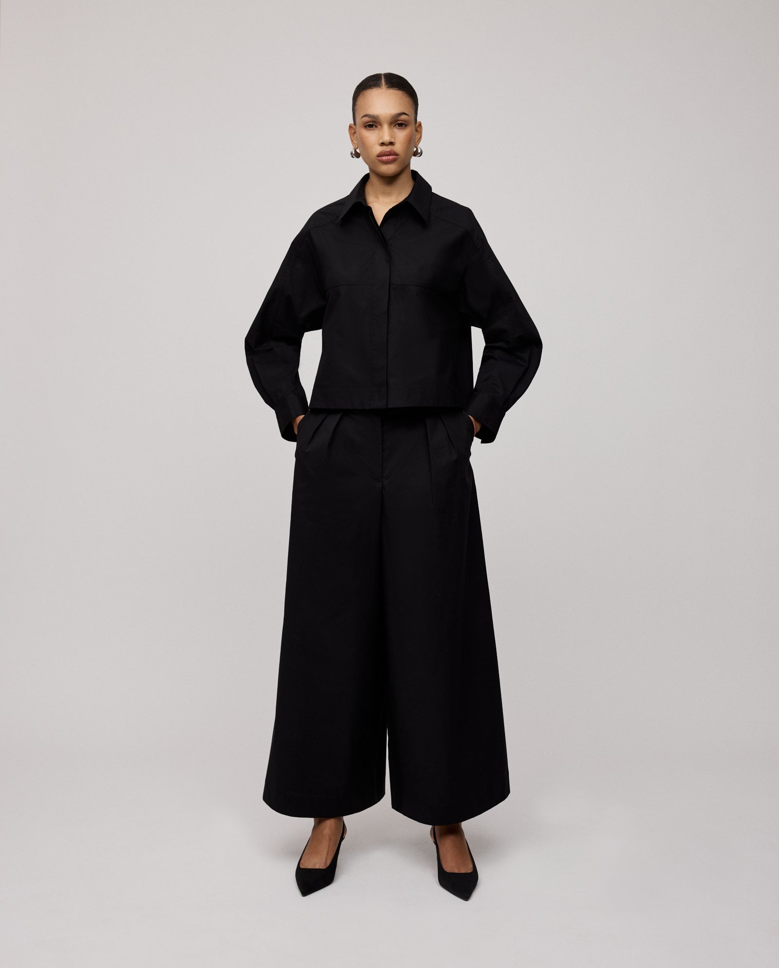 A woman stands with hands in pockets against a plain background, wearing IVY OAKs PIERINA Pants and a black collared jacket.