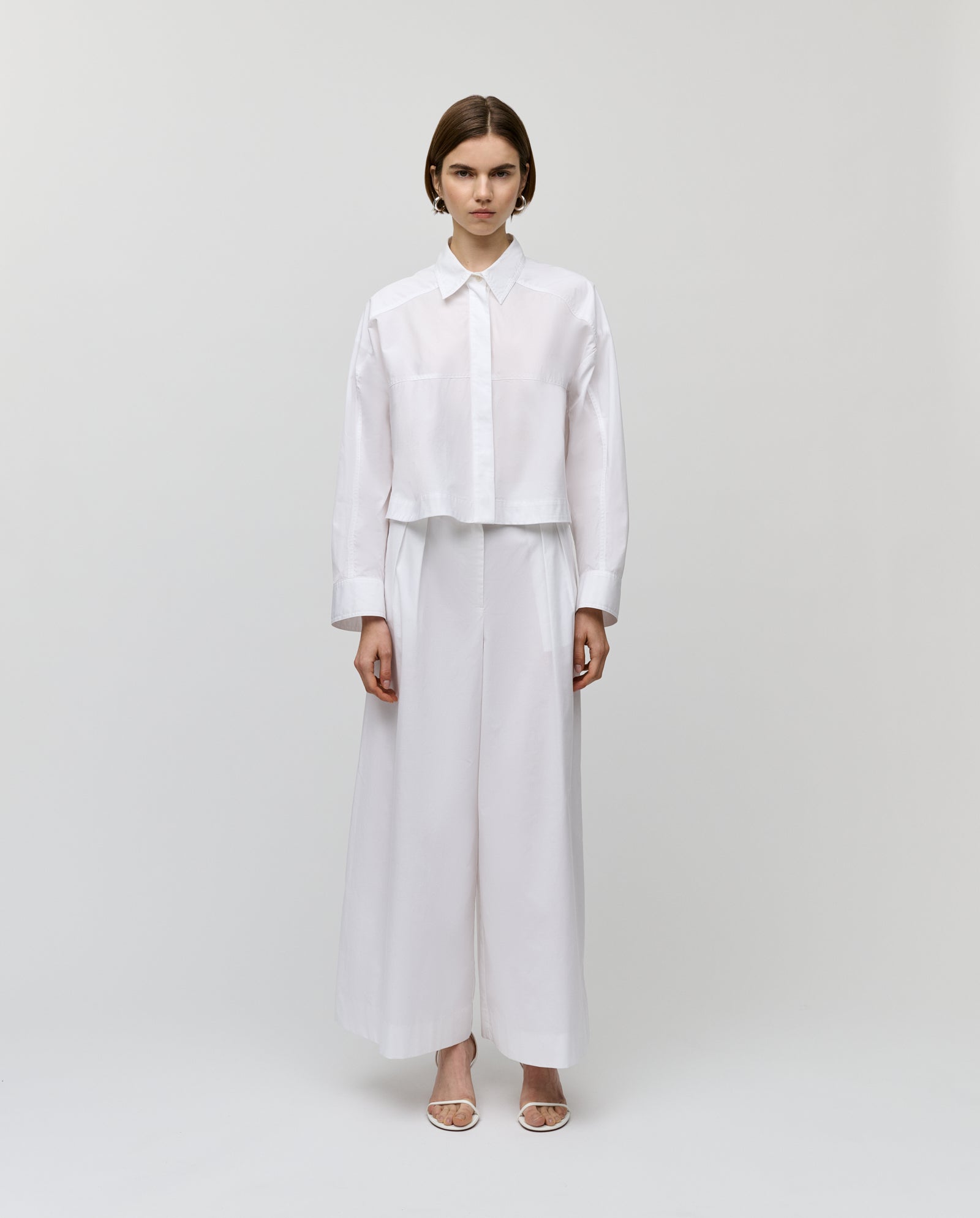 A woman wears IVY OAKs PIERINA Pants—long, wide-leg cotton trousers with subtle waist darts—paired with a white shirt, set against a plain white background.