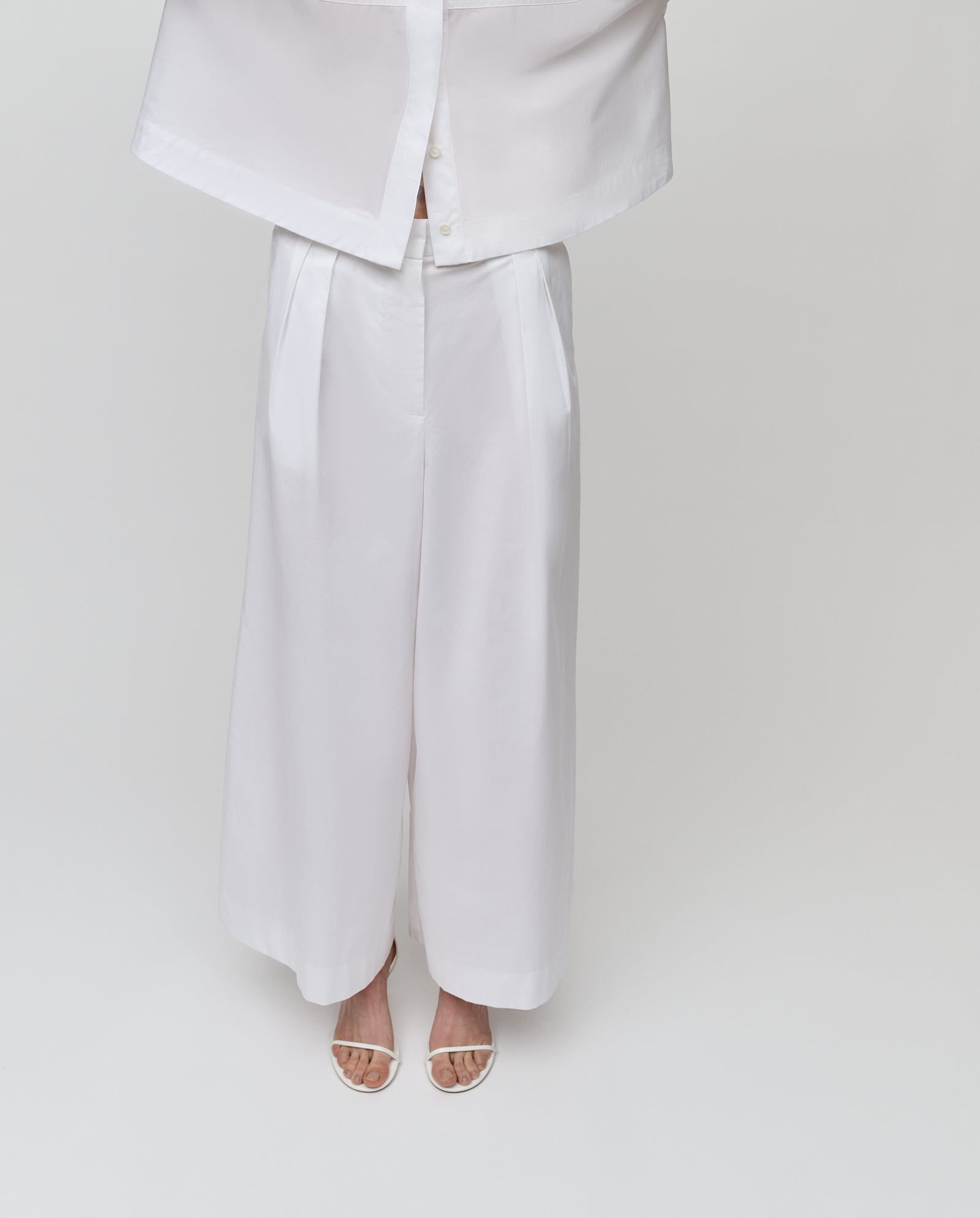 Model wears IVY OAKs PIERINA Pants, featuring a wide-leg cotton design with waist darts, paired with a matching white top and sandals, posed against a plain white background.