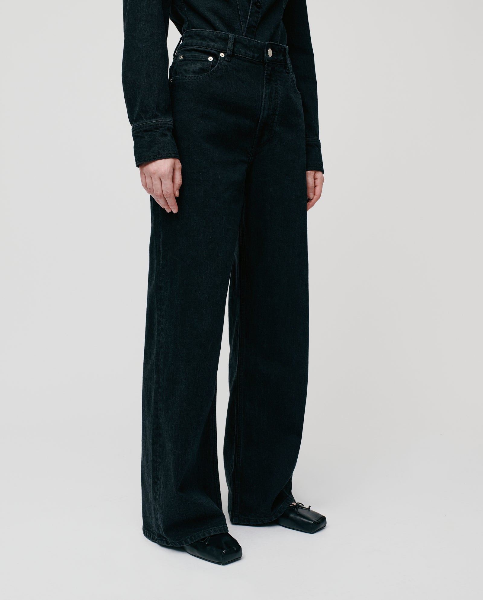 Model wears IVY OAK PRIJANKA high-waisted wide leg jeans in black, paired with a black long-sleeve shirt and black sandals, set against a plain background.