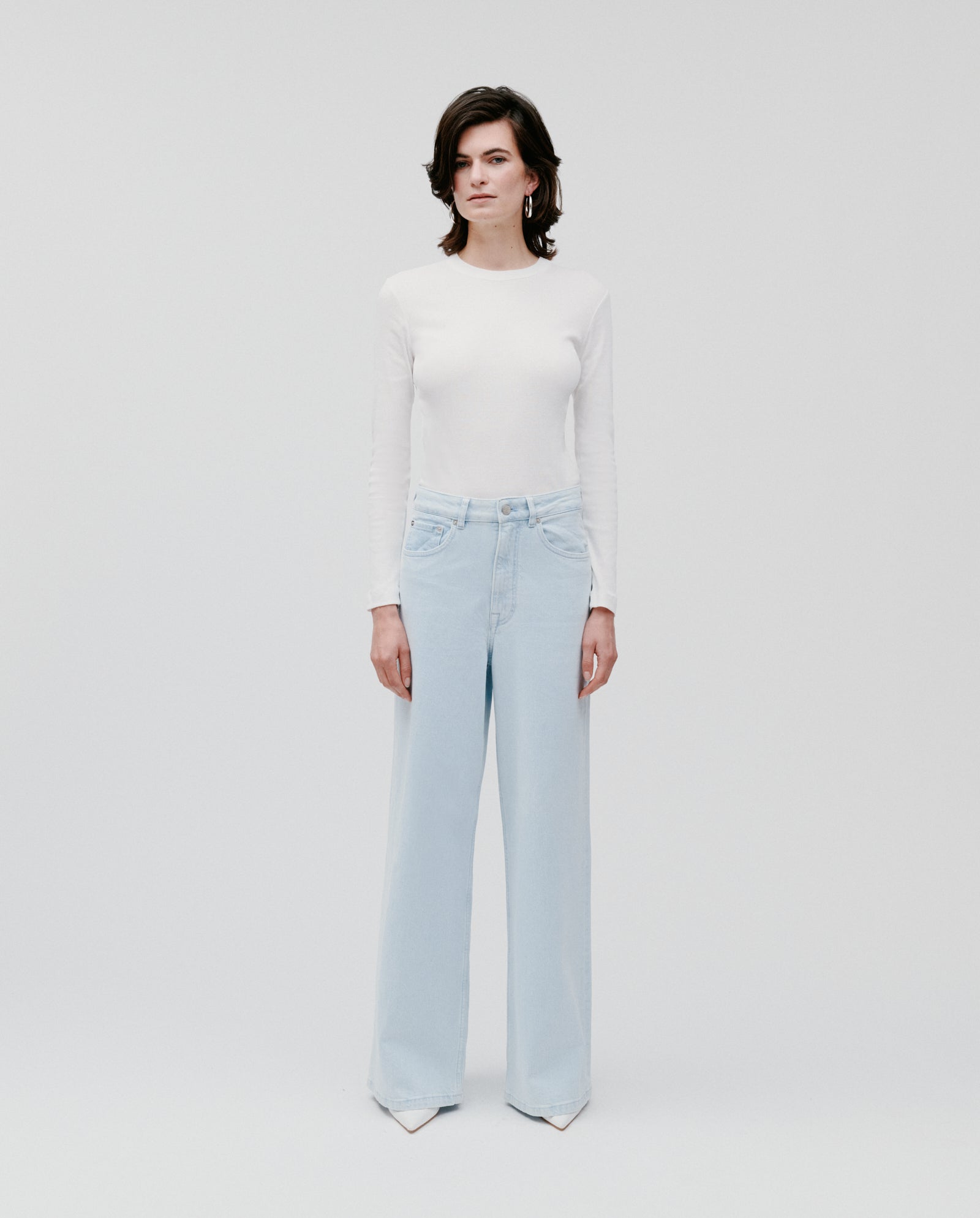 A person wearing a white long-sleeve shirt and IVY OAK PRIJANKA light blue high-waisted wide leg jeans stands against a plain white background.