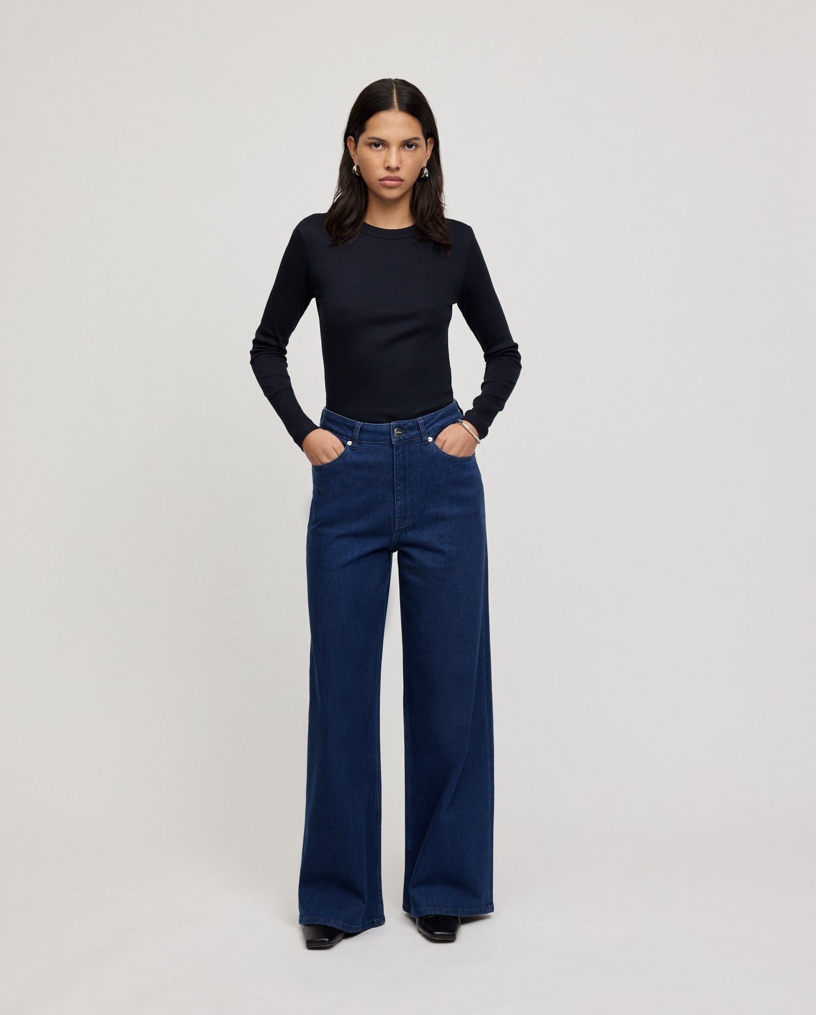 A woman stands with hands in pockets, wearing a black long-sleeve top and IVY OAK PRIJANKA Jeans—high-waisted, indigo denim with a loose, wide-leg silhouette.