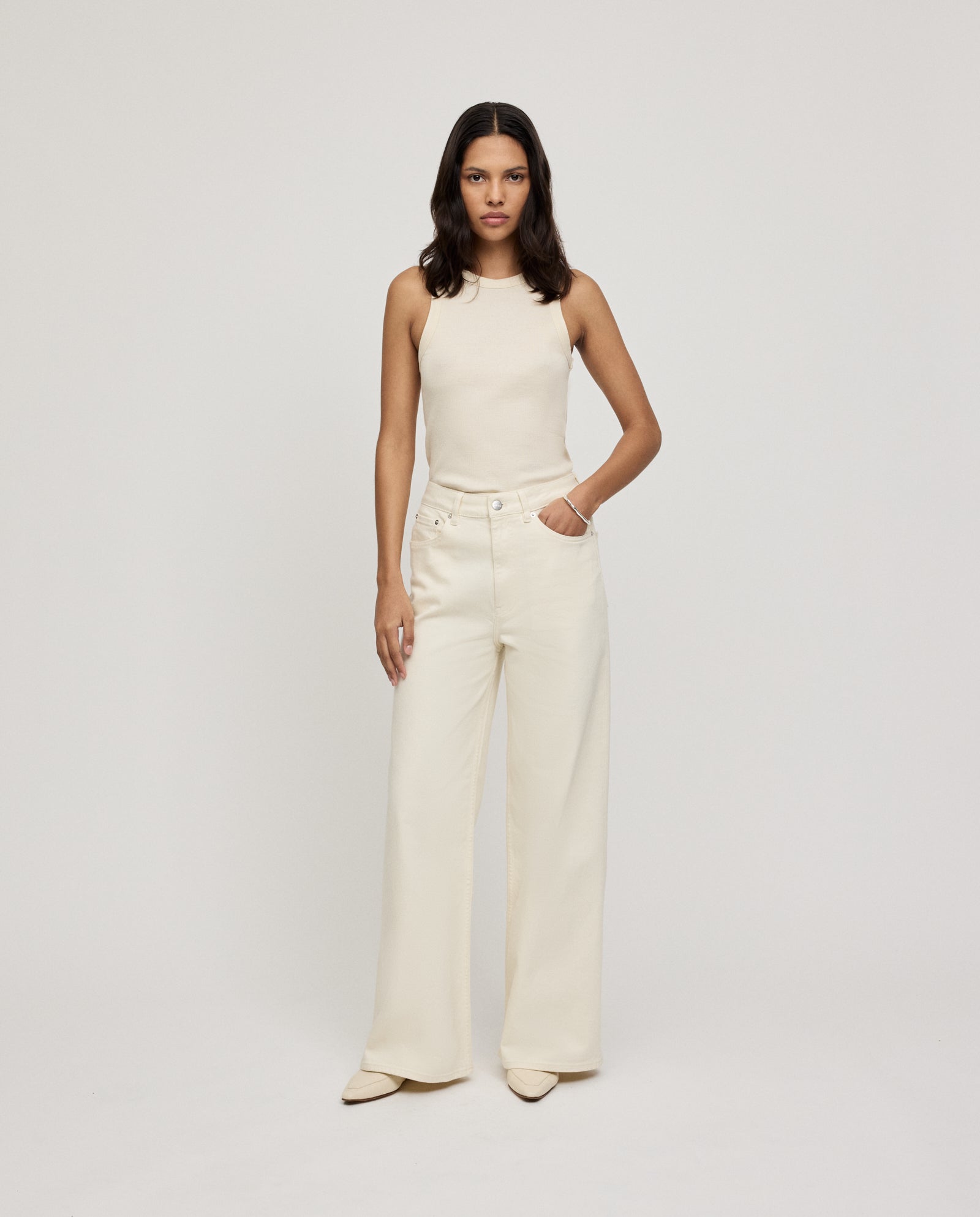 A woman wears high-waisted, loose wide-leg PRIJANKA Jeans by IVY OAK with a sleeveless cream top, standing with one hand in her pocket.