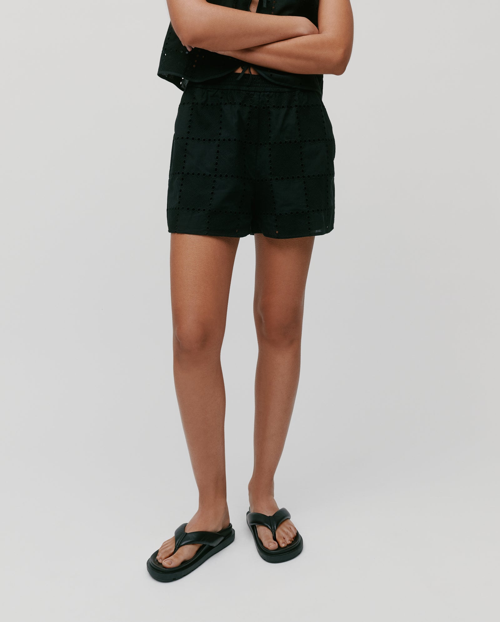 Person stands with arms crossed in black IVY OAK PIPER SUE Trousers featuring an elastic waistband, paired with a black top and black sandals, set against a plain background.