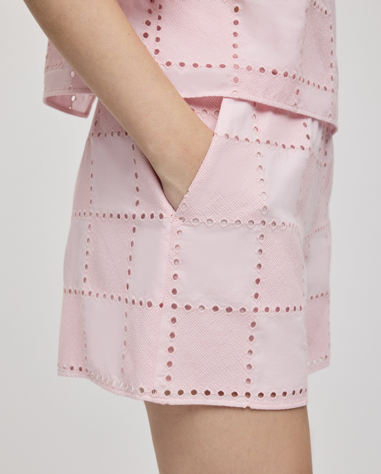 A person wears the IVY OAK PIPER SUE Trousers in light pink with broderie anglaise square patterns and an elasticated waist, paired with a matching top, hand in pocket.