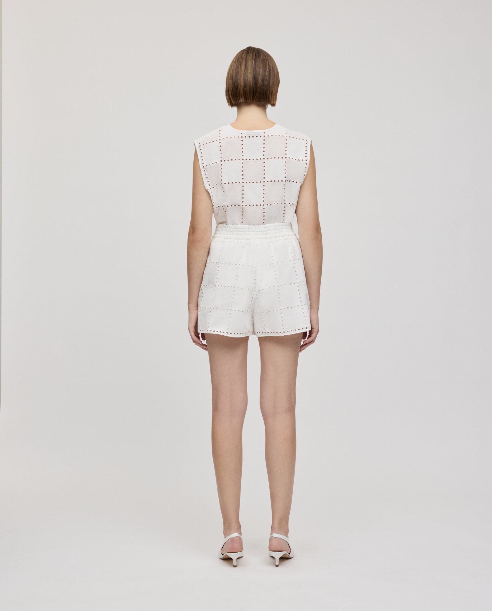 A woman stands with her back to the camera, wearing a sleeveless white top and IVY OAKs PIPER SUE Trousers, which feature an elastic waistband and a broderie anglaise cut-out square pattern.
