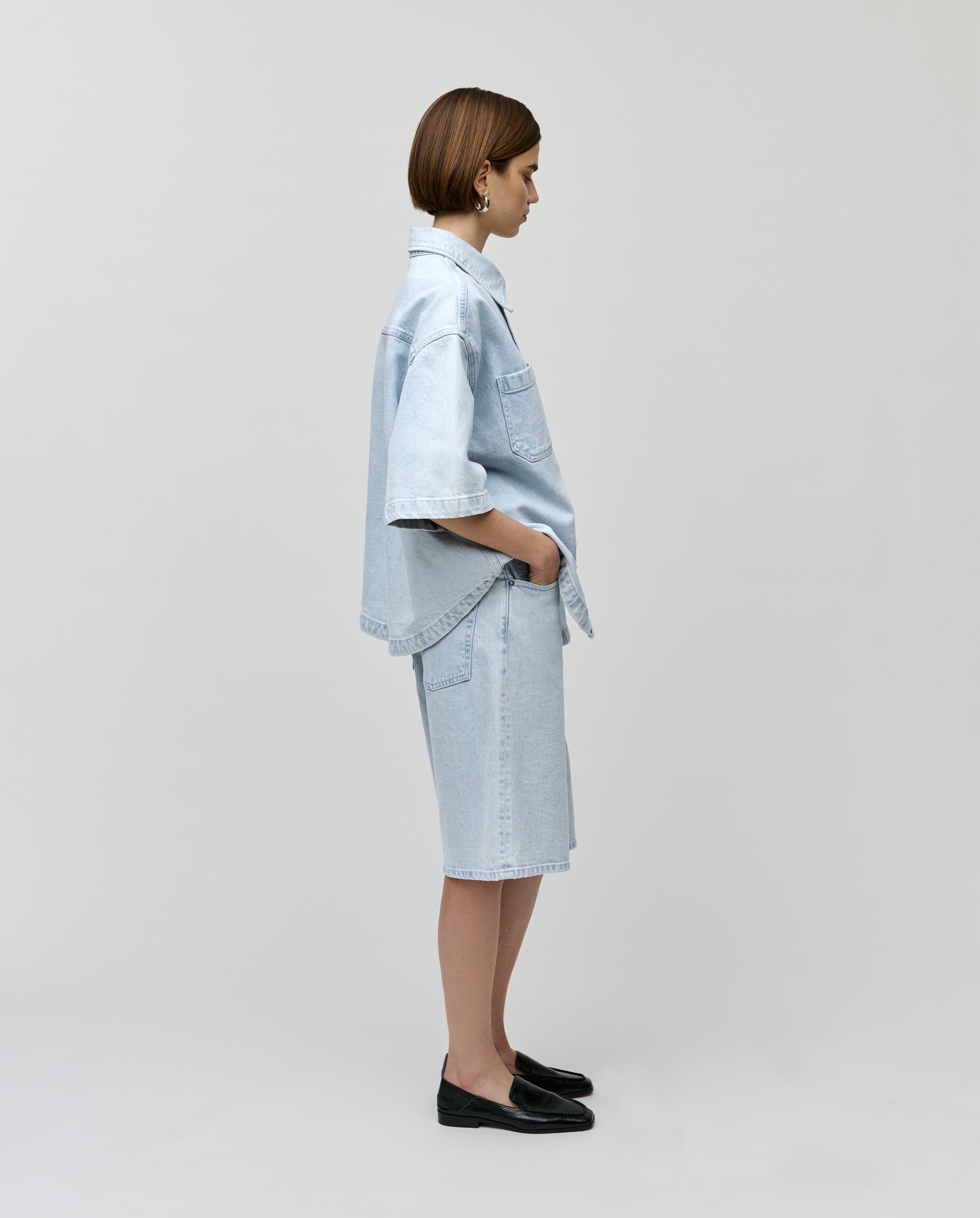 Woman standing in profile against a plain background, hands in pockets, wearing IVY OAKs PIRI Pants with a light denim shirt and classic jeans style.