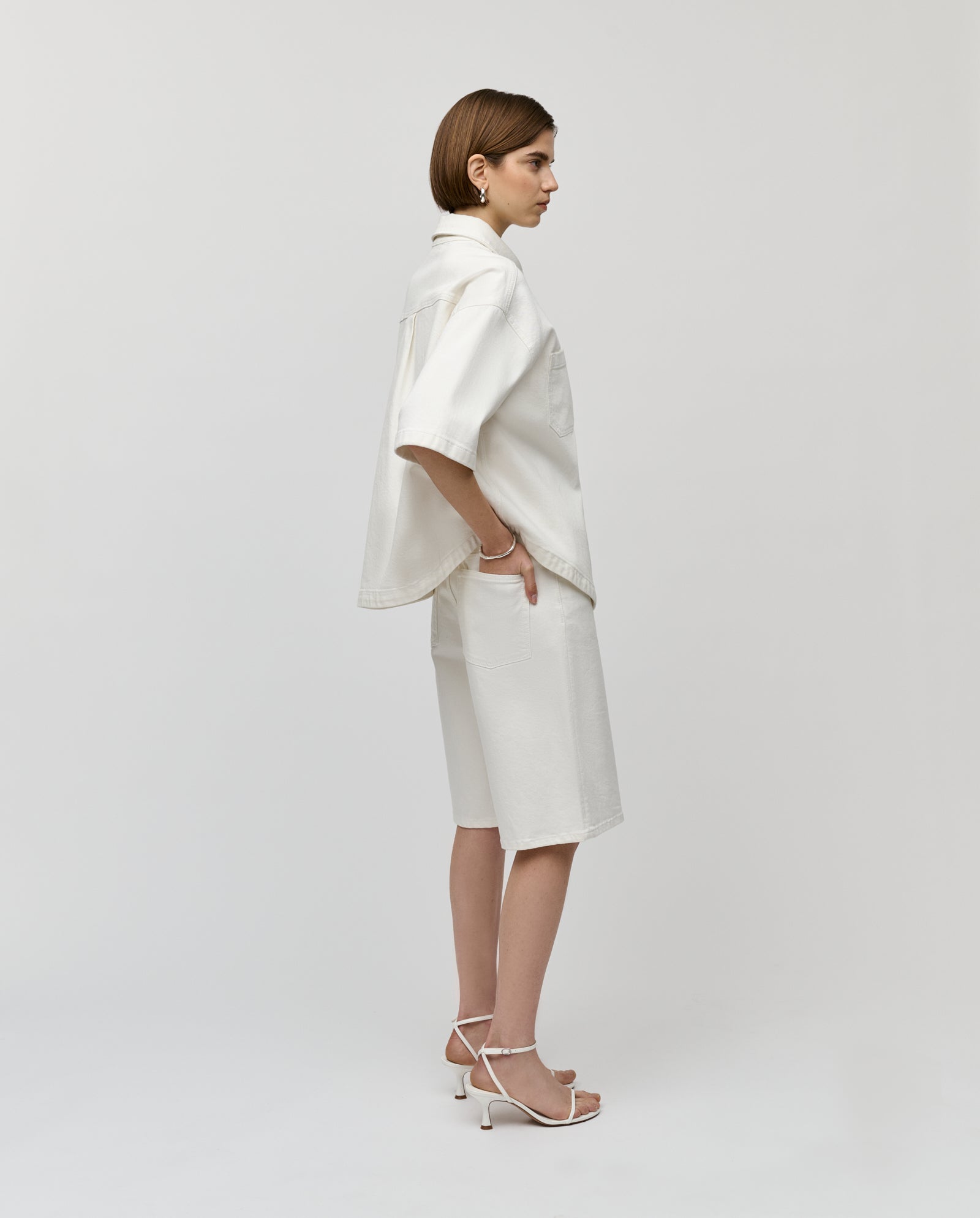 Woman wearing IVY OAK’s PIRI Pants and a white oversized shirt, standing sideways in white heels against a plain background.