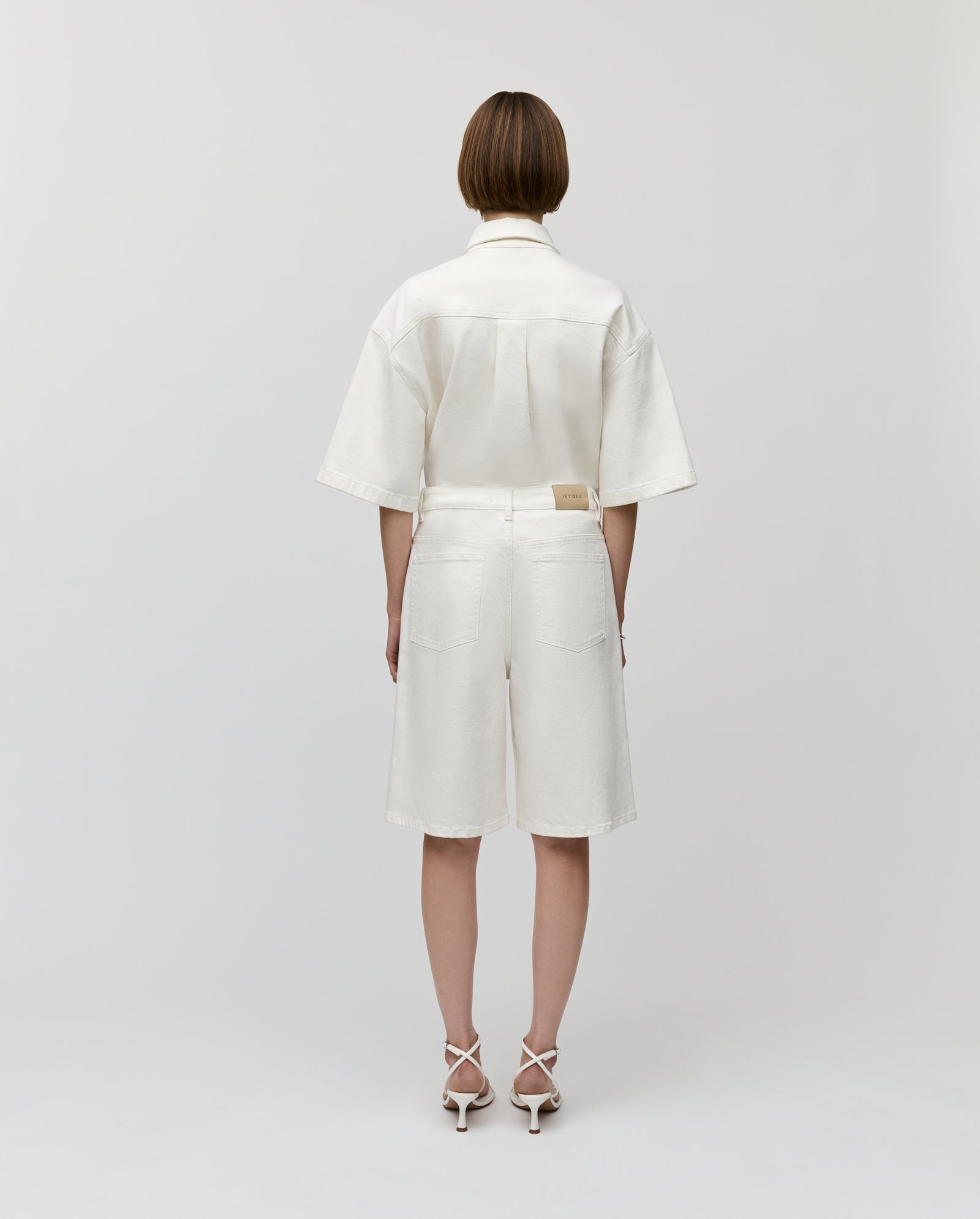 A woman stands with her back to the camera, wearing IVY OAKs PIRI Pants, a white shirt, and white heeled sandals.