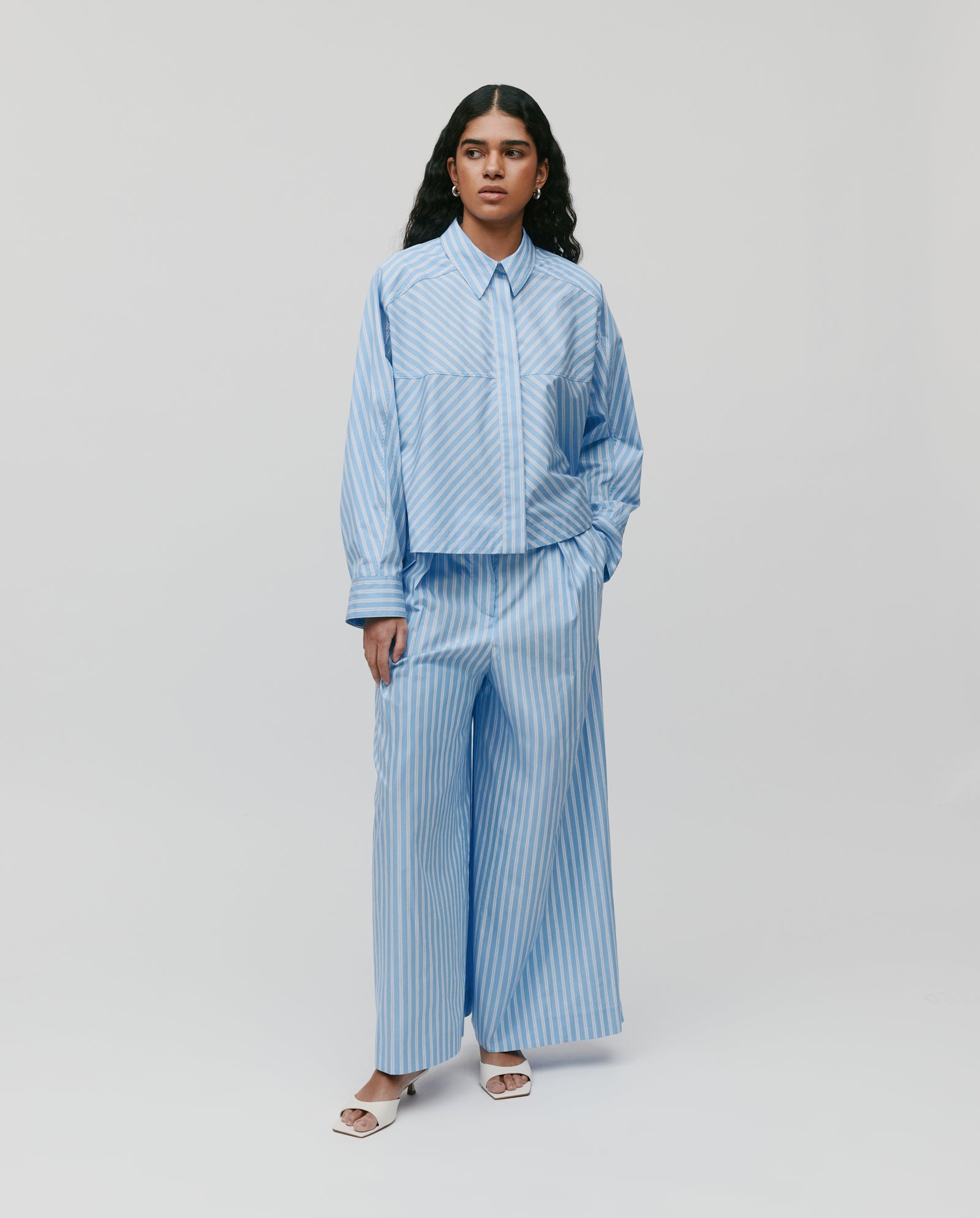 A woman stands against a white background wearing IVY OAKs PIERINA Pants—wide-leg, long cotton pants with waist darts—paired with a loose blue and white striped shirt.
