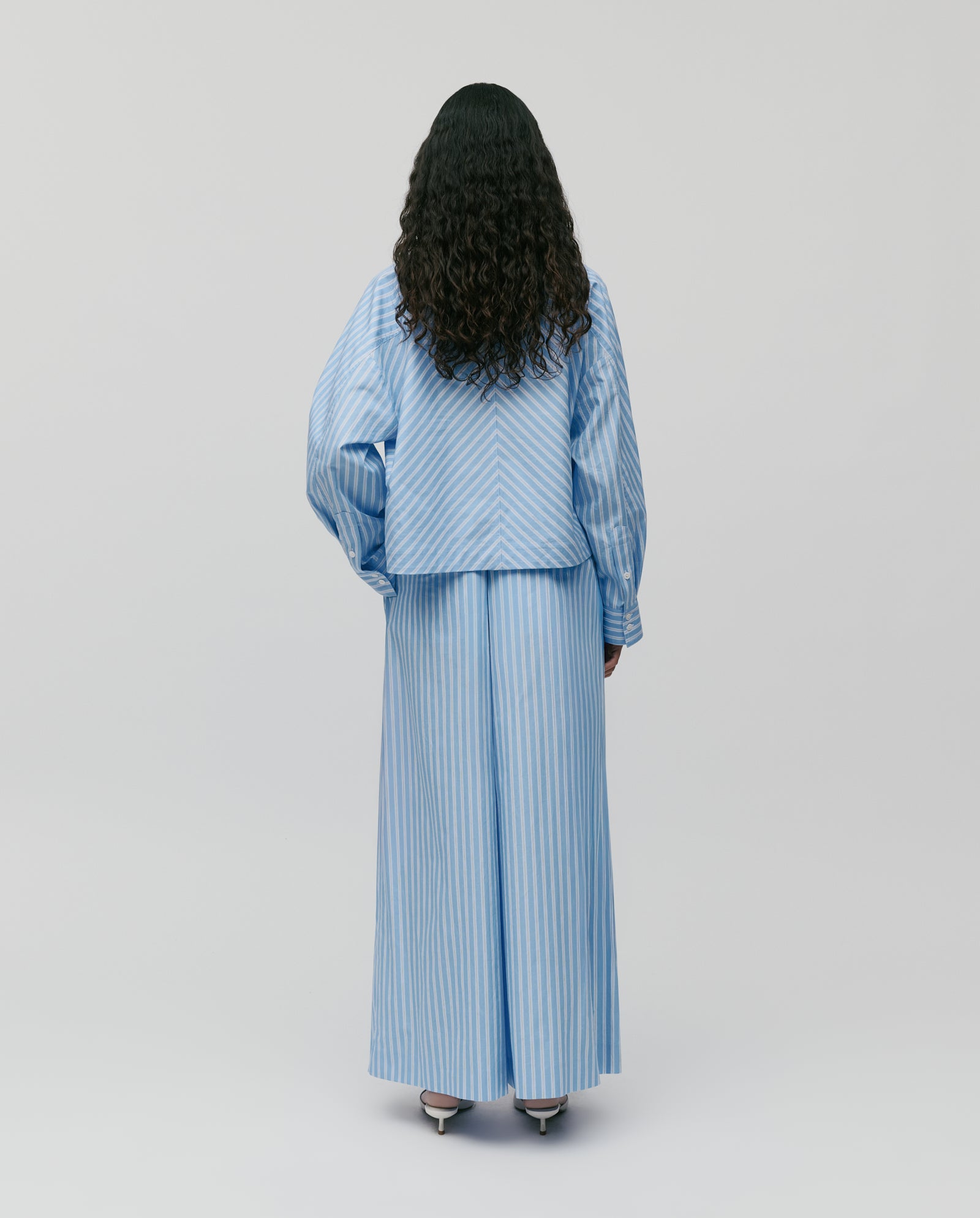 Person with long, curly hair stands with their back to the camera, wearing IVY OAK’s PIERINA Pants—wide leg cotton pants in a blue and white striped design.