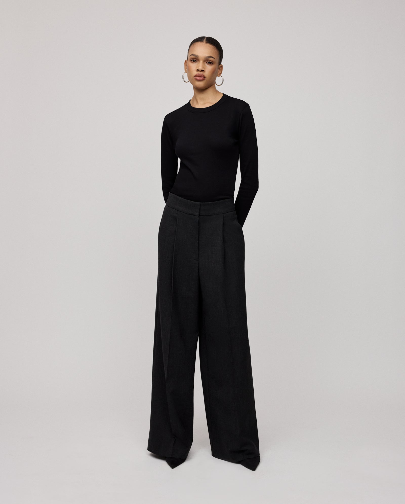 A woman stands with hands in pockets against a plain background, wearing a black long-sleeve top and IVY OAK PRESCILLIA SUE wool-blend twill wide leg pants.