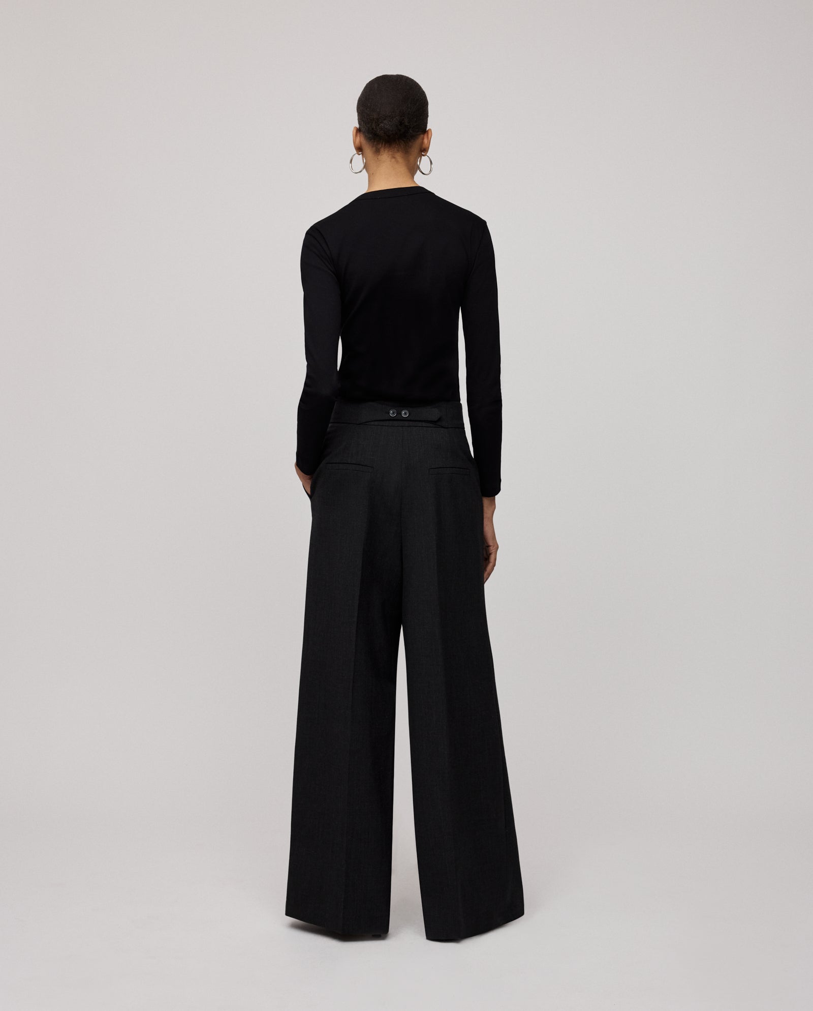 A person stands facing away against a plain background, wearing IVY OAKs PRESCILLIA SUE Pants and a black long-sleeve top.