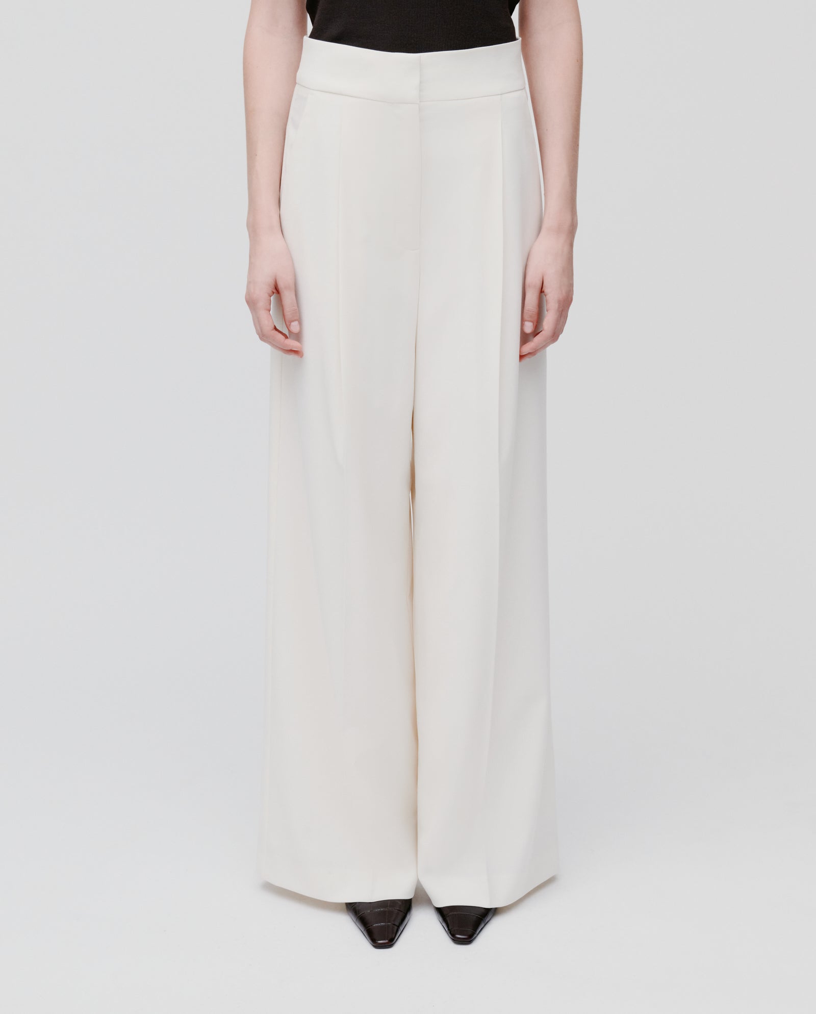 A model wears the IVY OAK PRESCILLIA SUE Pants, featuring a white wide-leg silhouette, styled with a black top and black pointed shoes against a plain background.