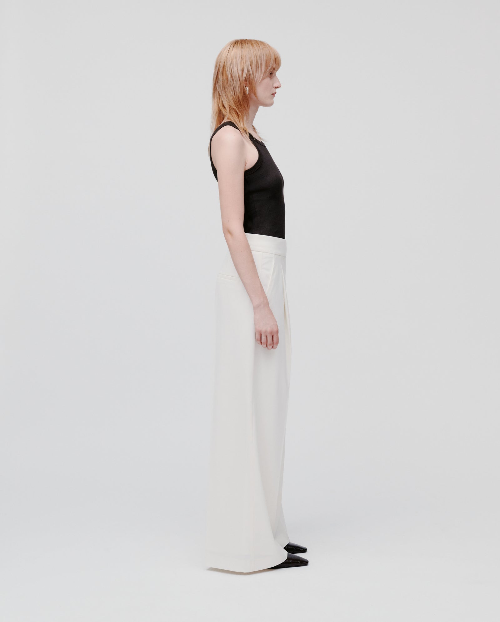 A person stands in profile against a plain background, wearing IVY OAKs PRESCILLIA SUE Pants in white and a black tank top.