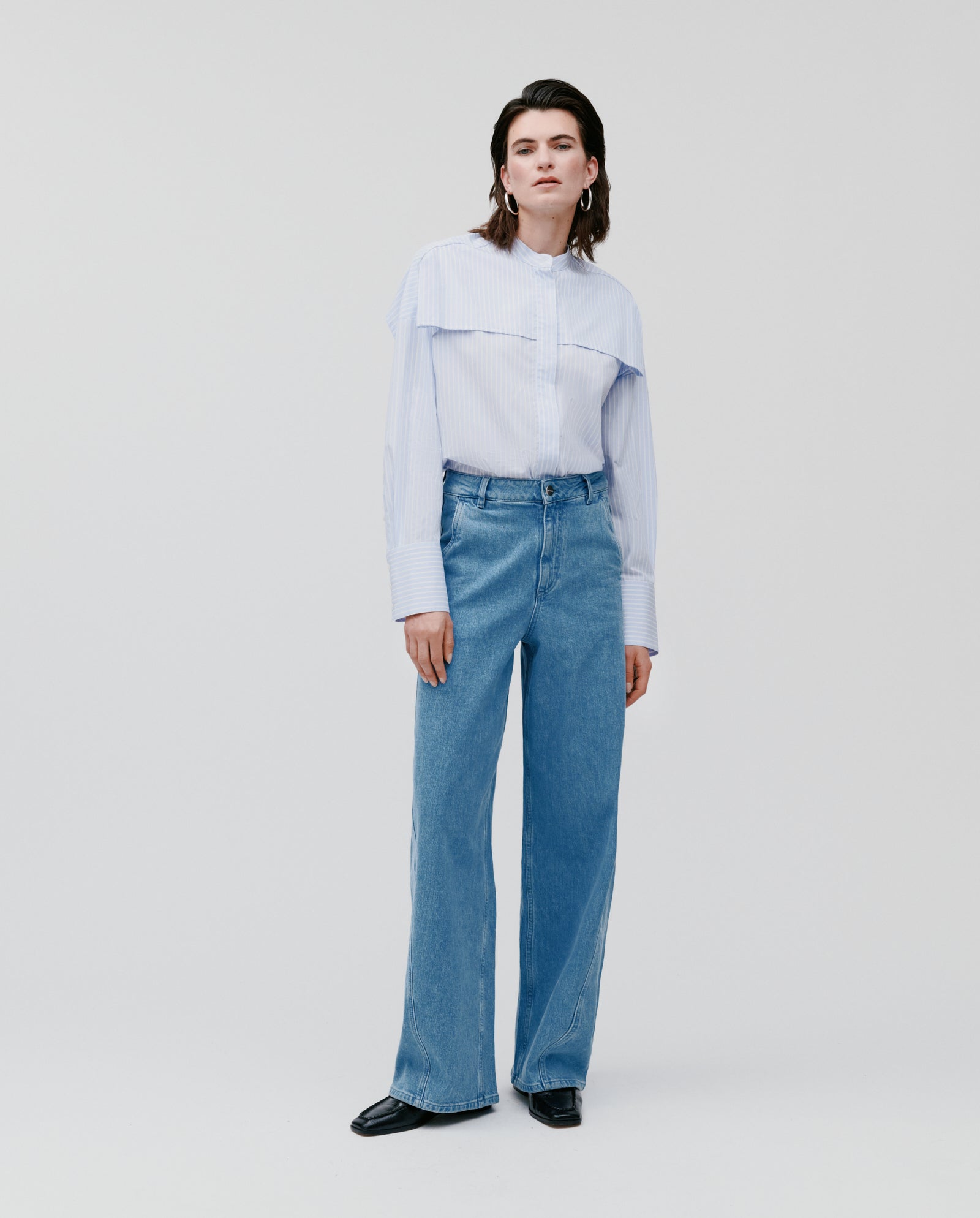 A person stands against a white background, wearing a light blue collared shirt and IVY OAKs PHILA Pants for women, featuring a barrel leg silhouette.