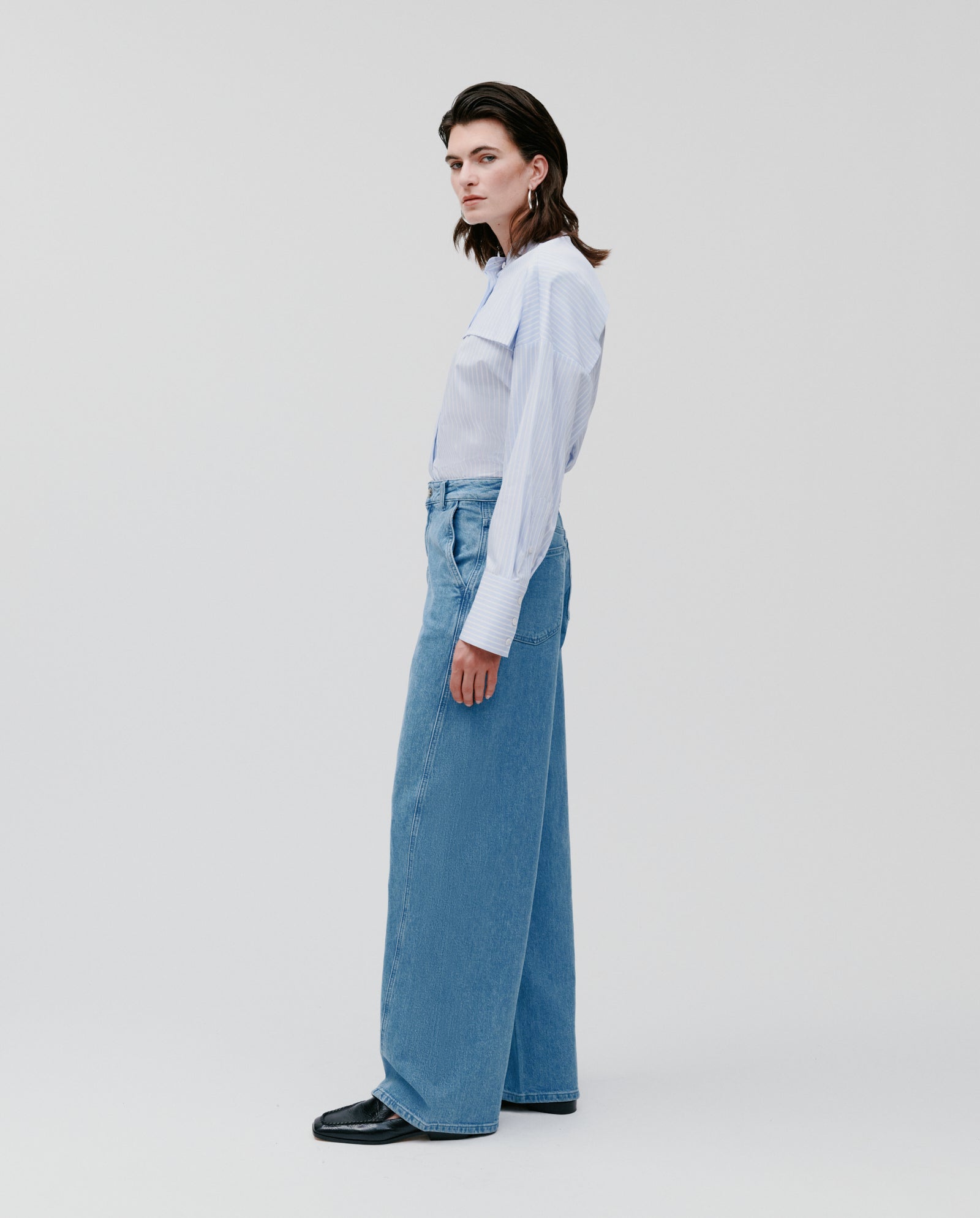 A person stands sideways against a plain background, wearing a light blue shirt and IVY OAKs PHILA Pants—deconstructed denim barrel leg style.