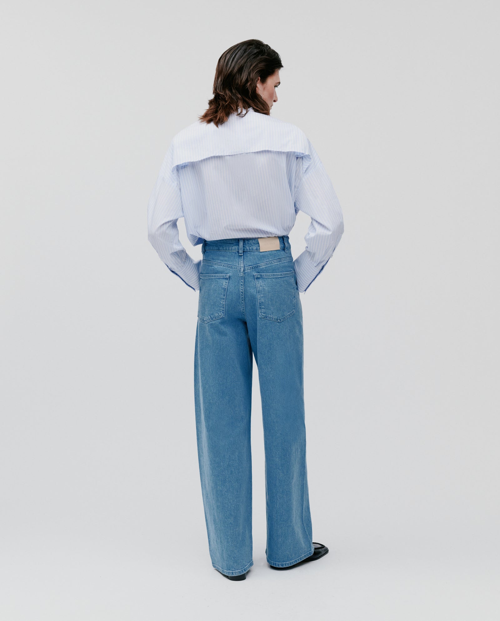 A person stands with their back to the camera, wearing a striped shirt and IVY OAK PHILA Pants—women’s deconstructed denim trousers with wide-barrel legs—against a plain background.