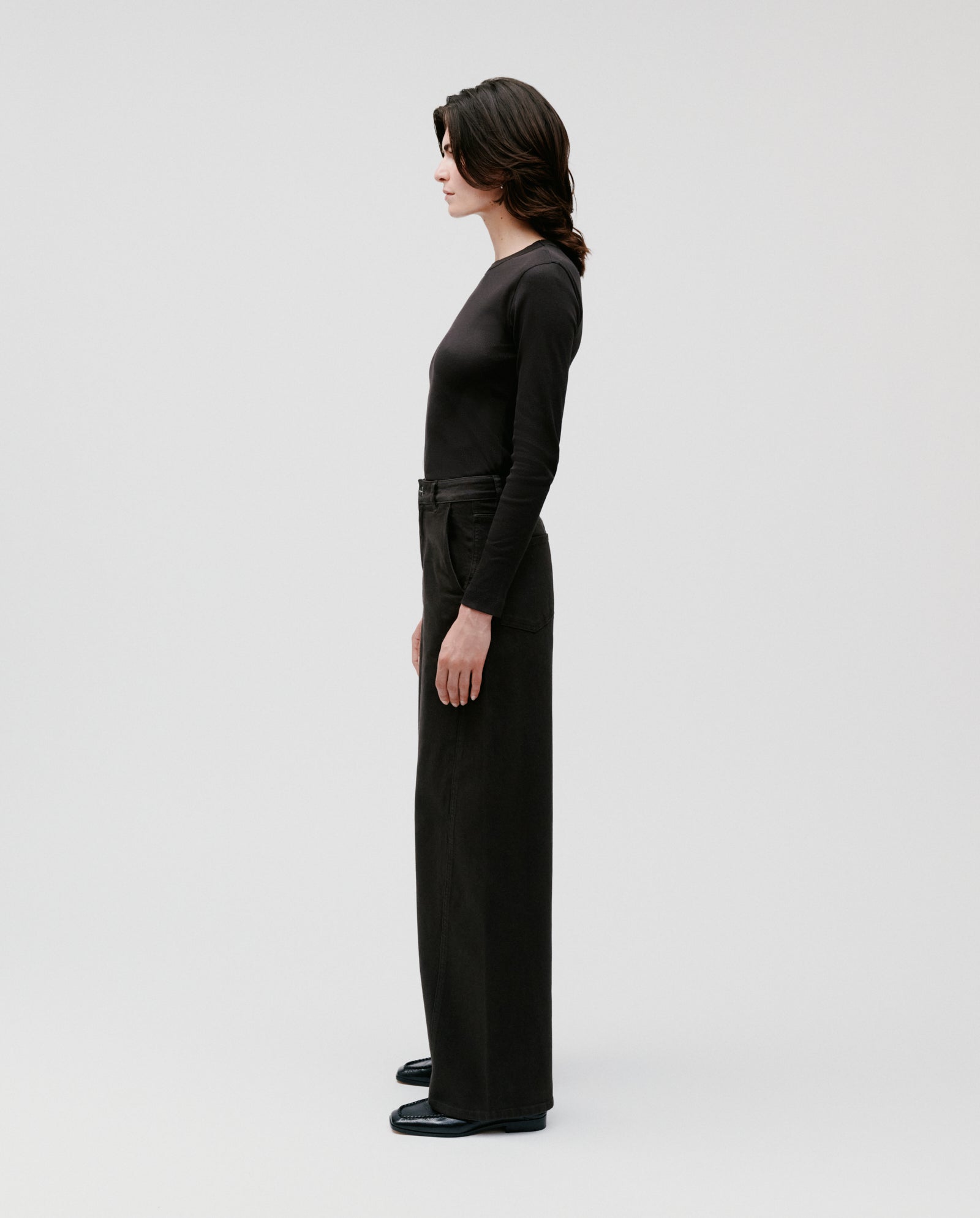 Wearing IVY OAK’s PHILA Pants and a black long-sleeve top, a woman stands in profile against a plain light background.